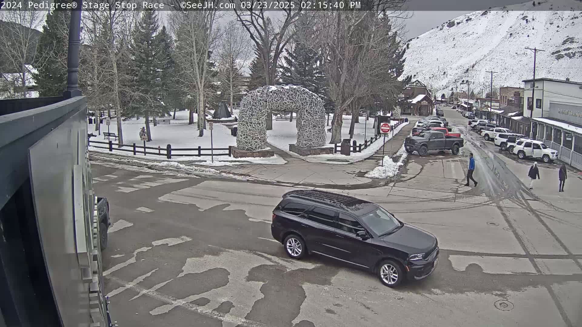 A black SUV is driving through a snow-covered town square with a large antler archway in the background under an overcast sky.