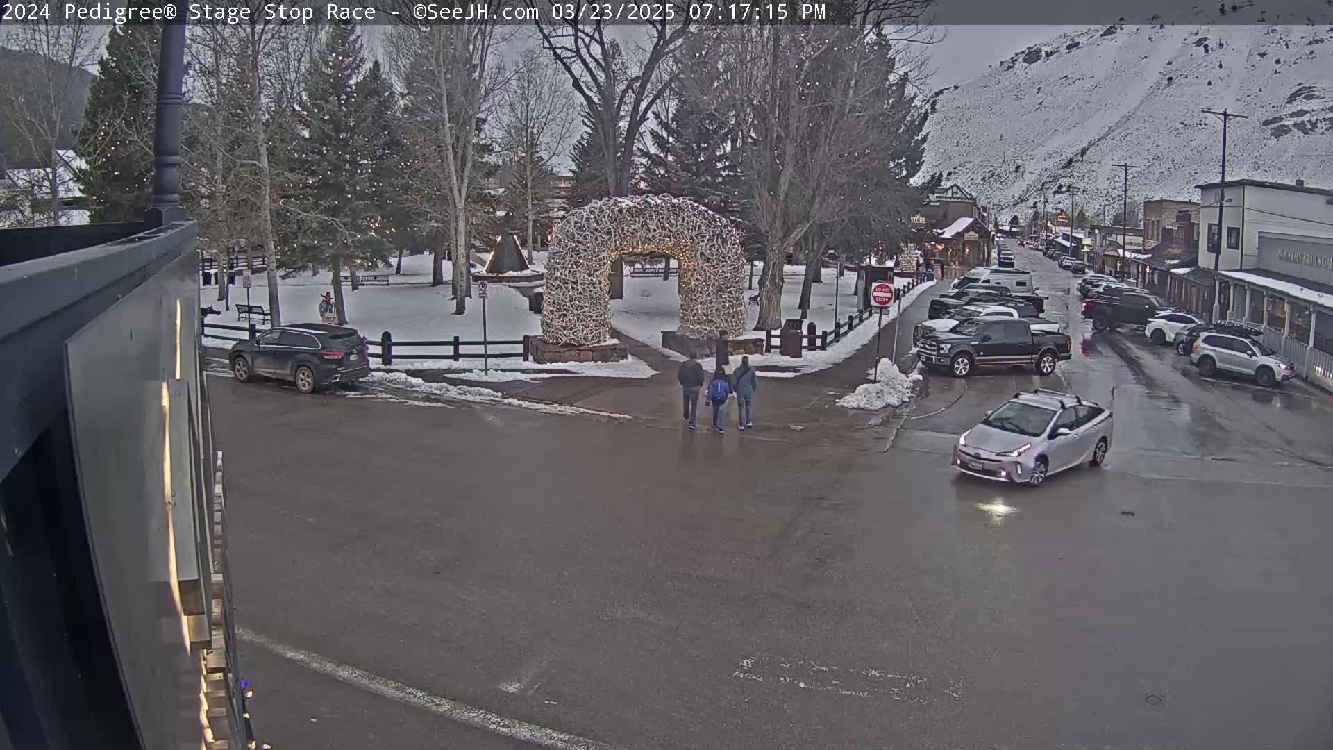 A wet street scene shows several cars parked and driving near a snow-covered town square with a large antler archway, and three people walking toward it.