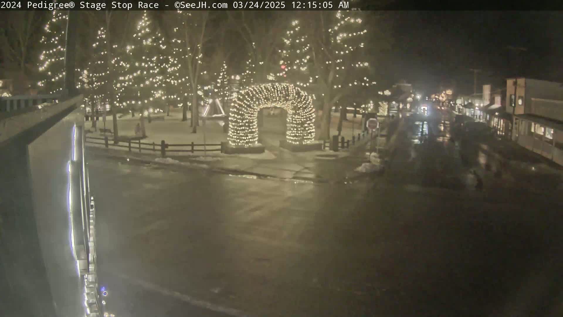 A nighttime view of a snow-covered town square with illuminated trees and a light-covered archway, wet roads, and a few buildings visible in the background.