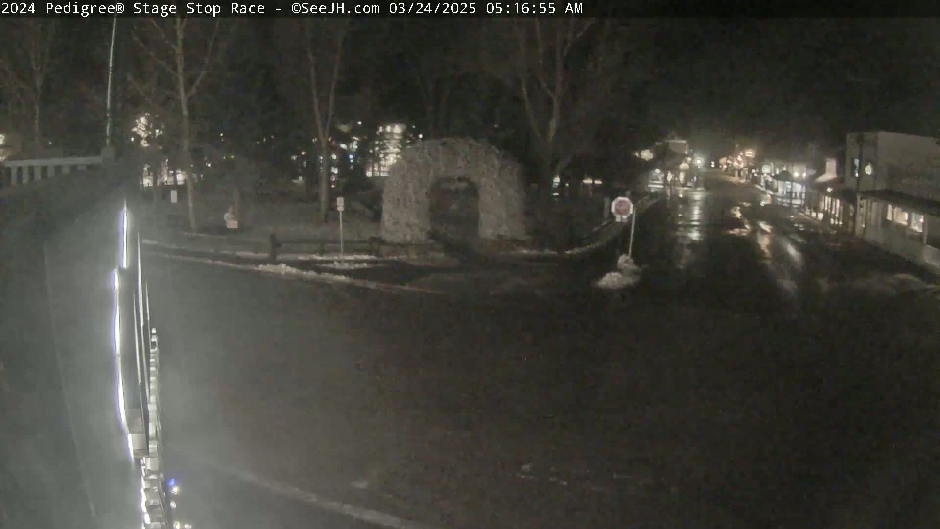 A nighttime, wet street scene shows a road with some buildings in the distance, a stone structure in the middle ground, and a bit of snow on the ground.