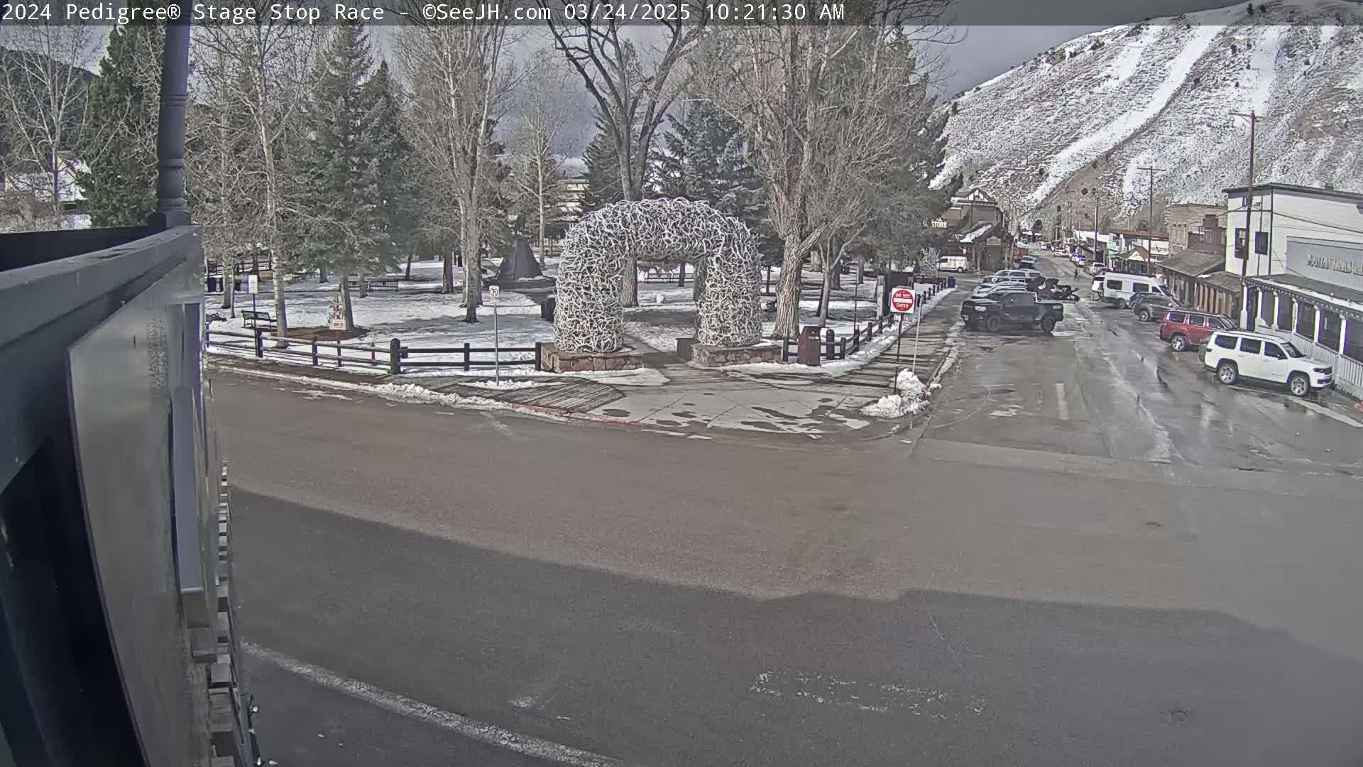 A snow-dusted town scene shows a street with parked cars, a small park with a large antler arch, and a snow-covered mountain in the background under an overcast sky.