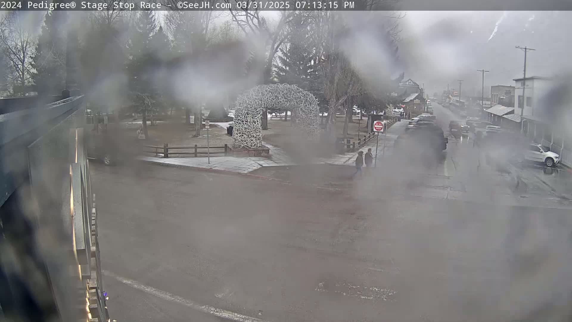 A rain-covered view of a town street shows a large antler arch in a park, several parked cars, and pedestrians walking near a crosswalk.