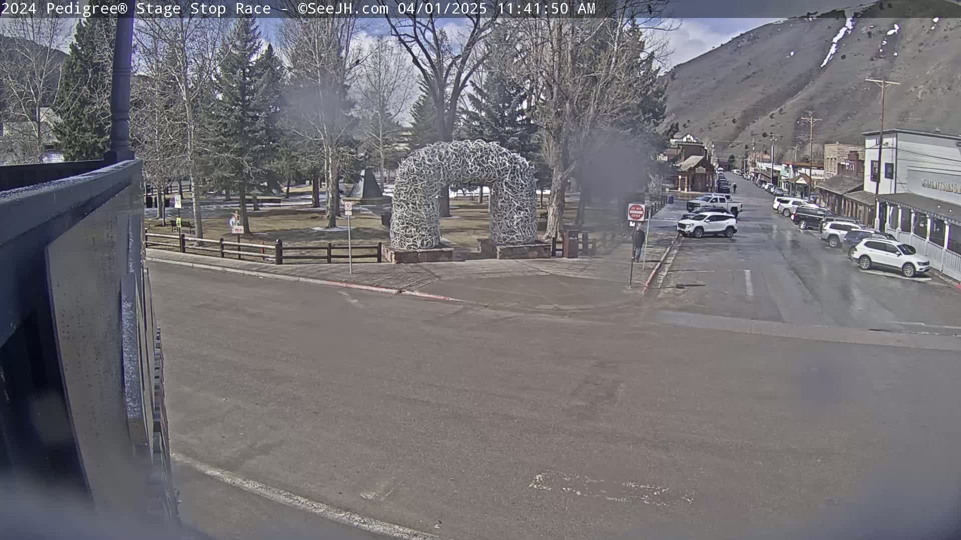 A partially visible structure is situated near a town square featuring a large antler arch, with several vehicles parked along a street next to buildings under a partly sunny sky.