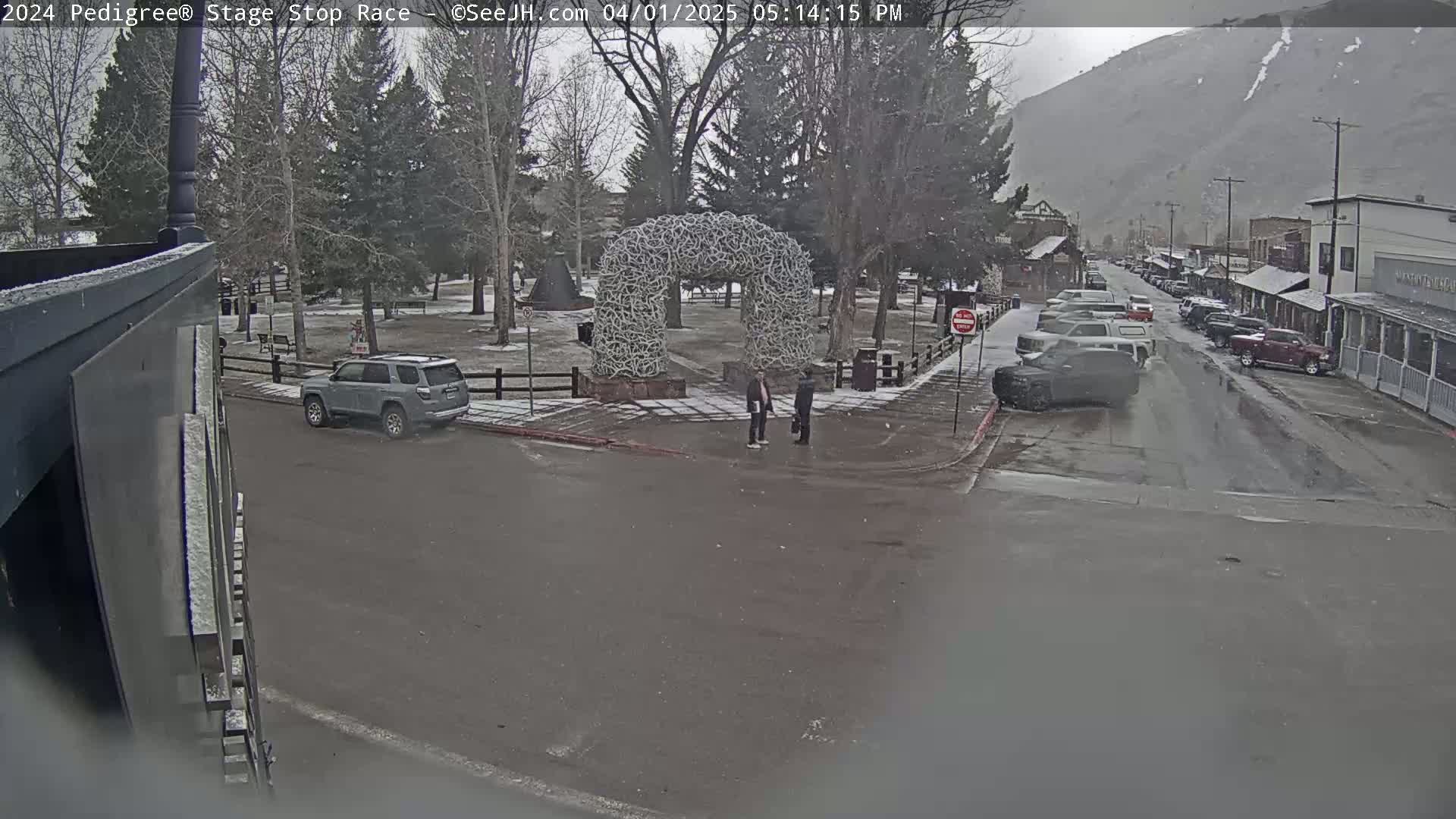 A snowy street scene shows a town square with a large antler arch, several parked cars, and two people standing near the arch, with a mountain visible in the background.