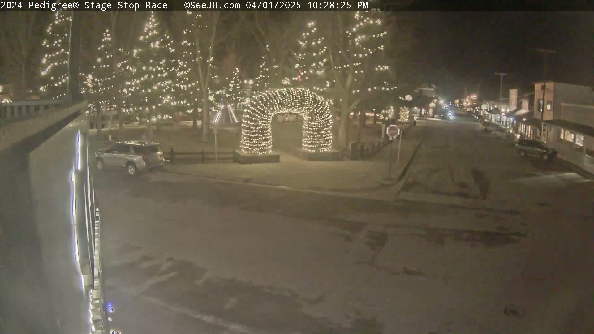 A snow-covered street at night, lined with Christmas-lit trees, features a light-up archway in the center and a few vehicles parked along the sides.