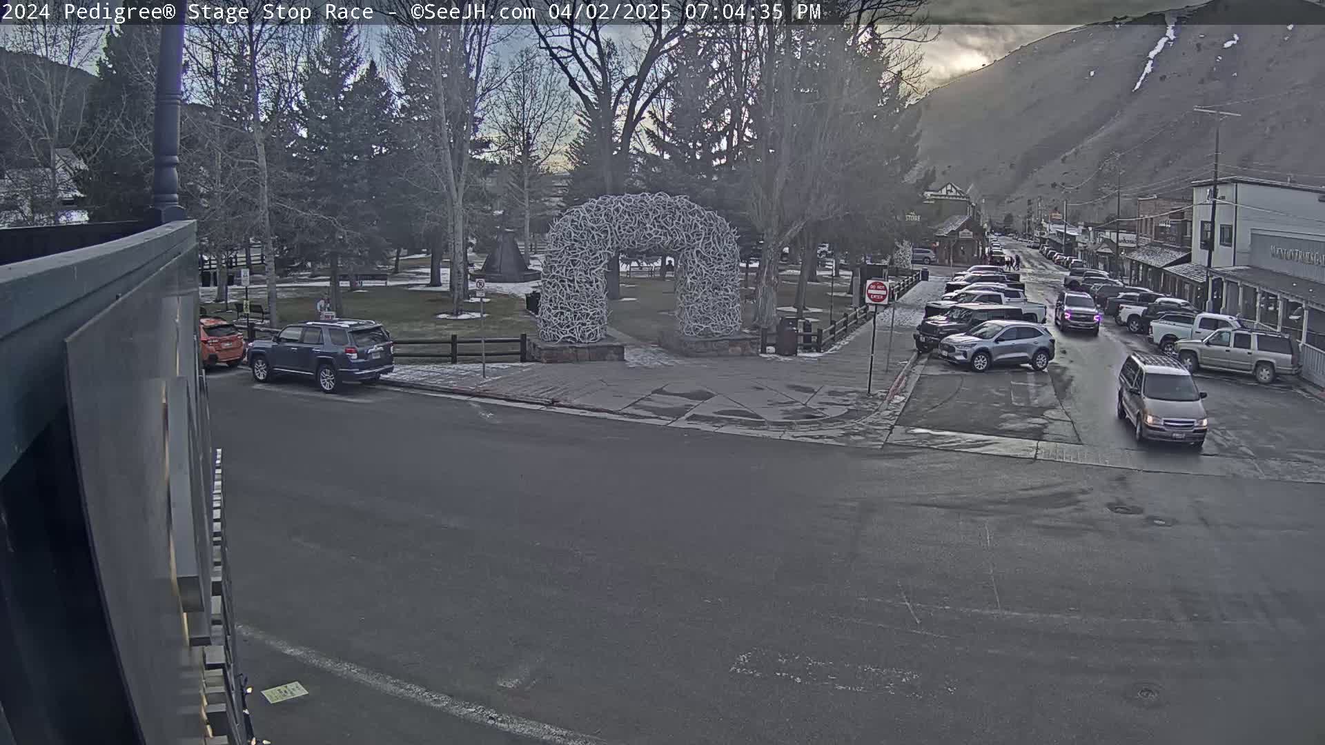 A town square with a large antler archway is seen on an overcast day, with several cars parked along the street and a few driving through the area.