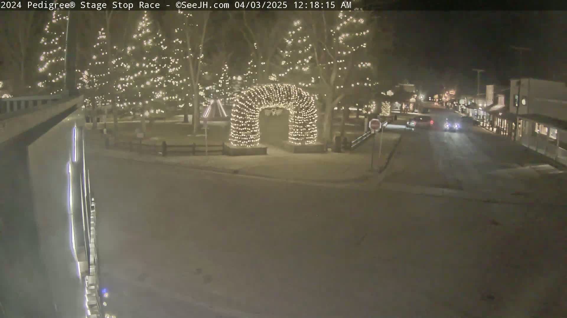 A snow-covered town square at night is illuminated by string lights on trees and an archway, with a few cars driving down a street.