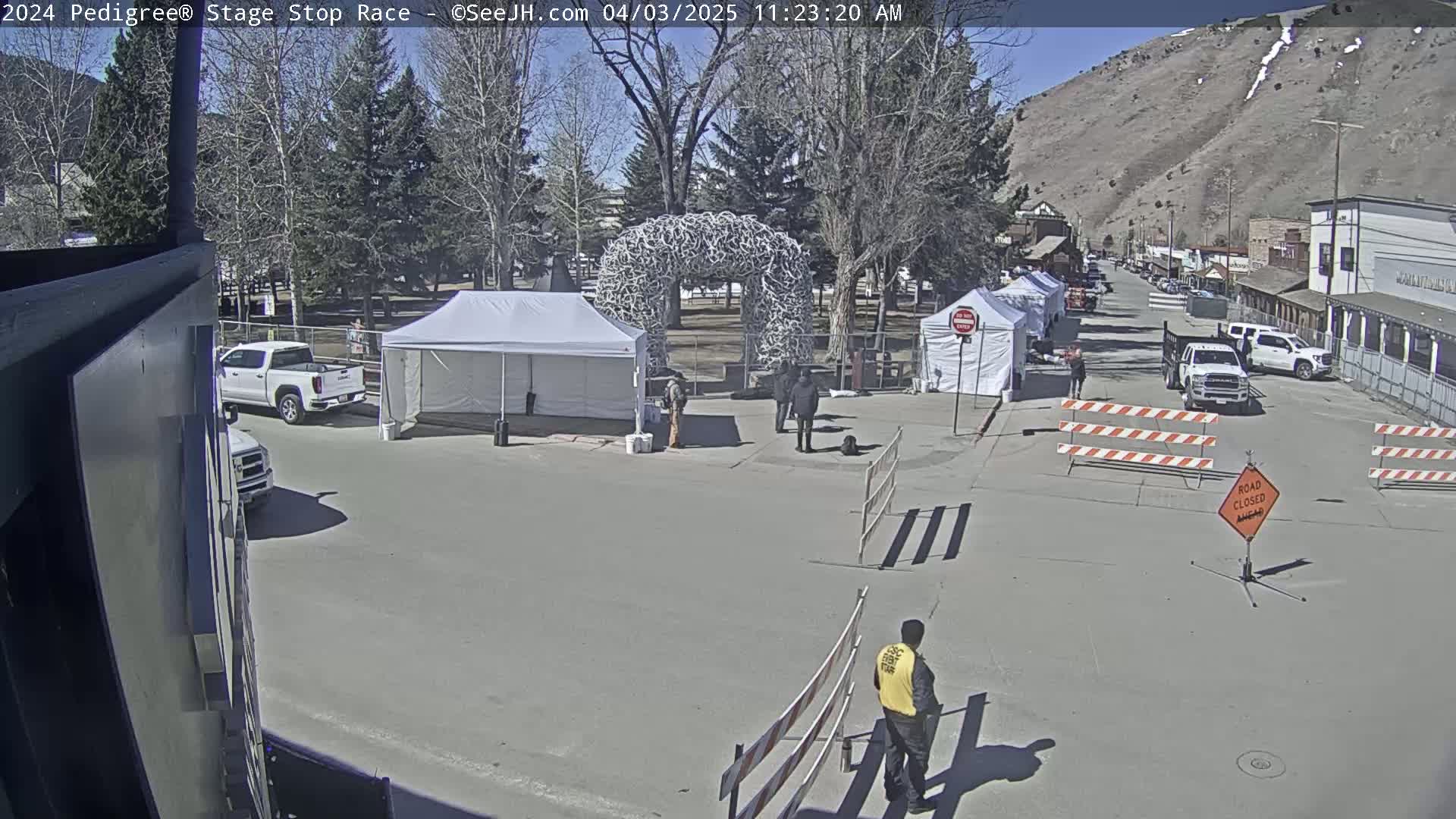 A sunny day reveals a town square with several tents, a large antler arch, parked vehicles, and road closure barriers, with a person in a safety vest standing near the barriers.