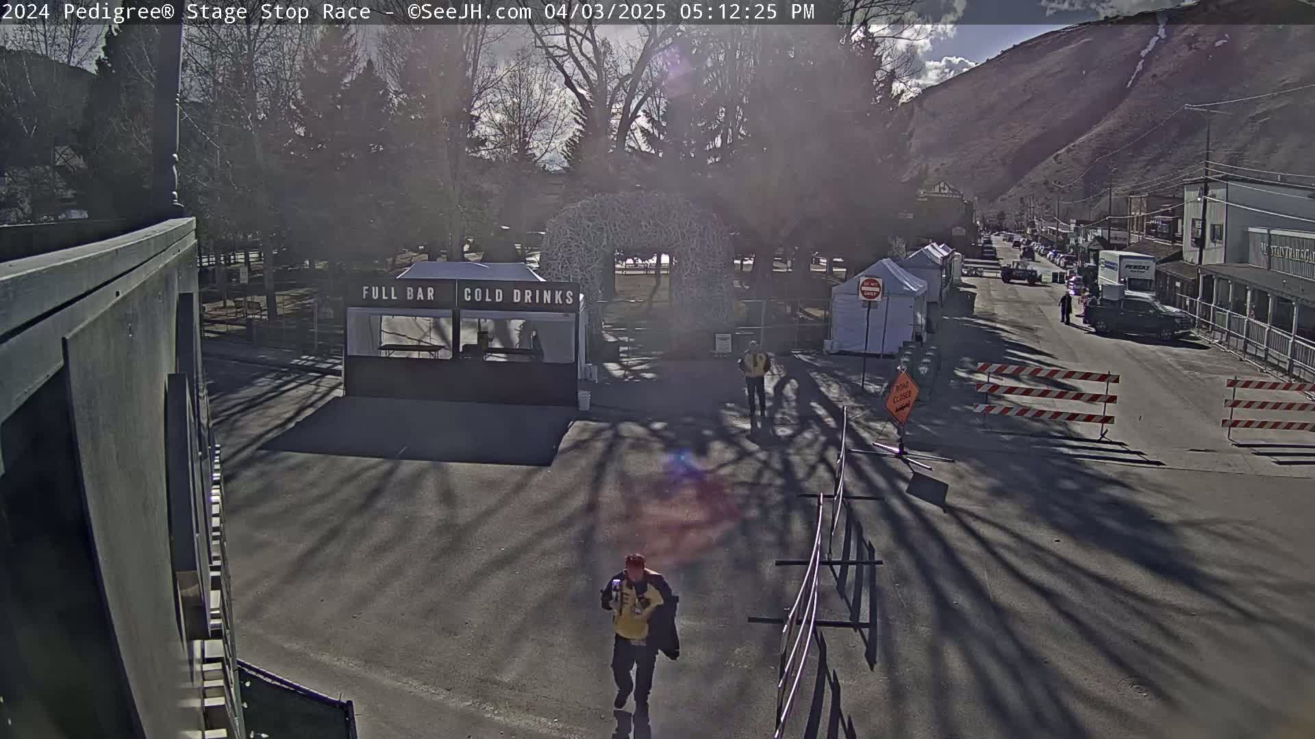 A sunny day reveals a mostly empty paved area with a few food and beverage tents, a person walking, and some traffic control barriers in front of buildings and a mountain in the background.