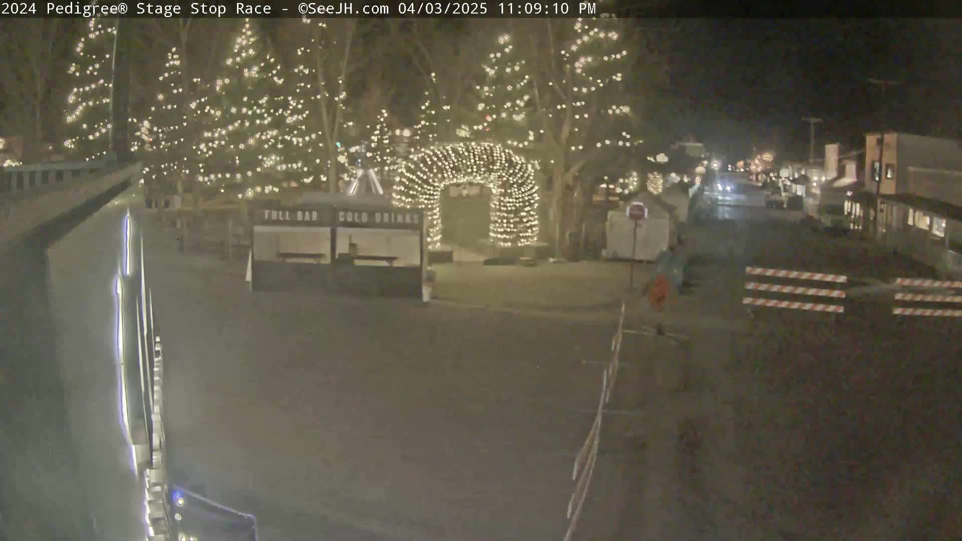A nighttime street scene shows a roadway with barriers, several buildings, and trees decorated with string lights.