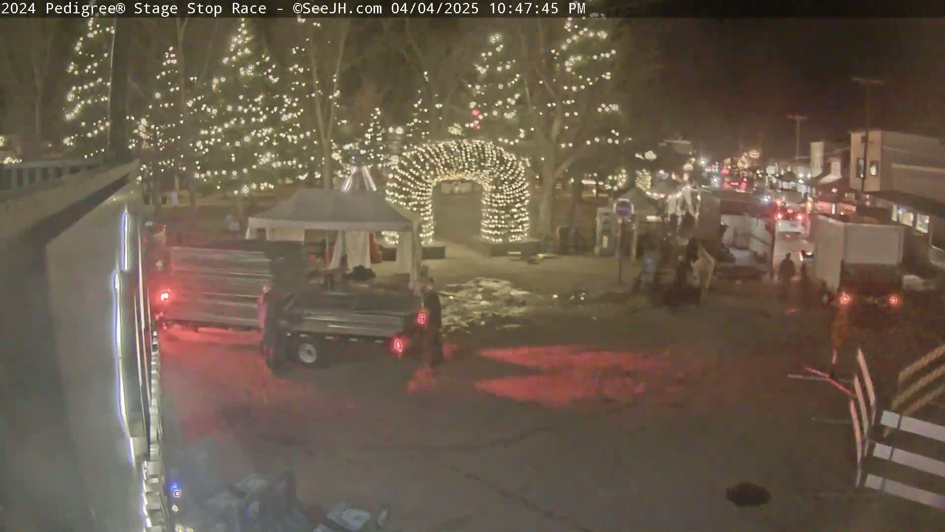 A nighttime scene shows a snowy town square with several trucks and people near a lighted archway and Christmas trees.