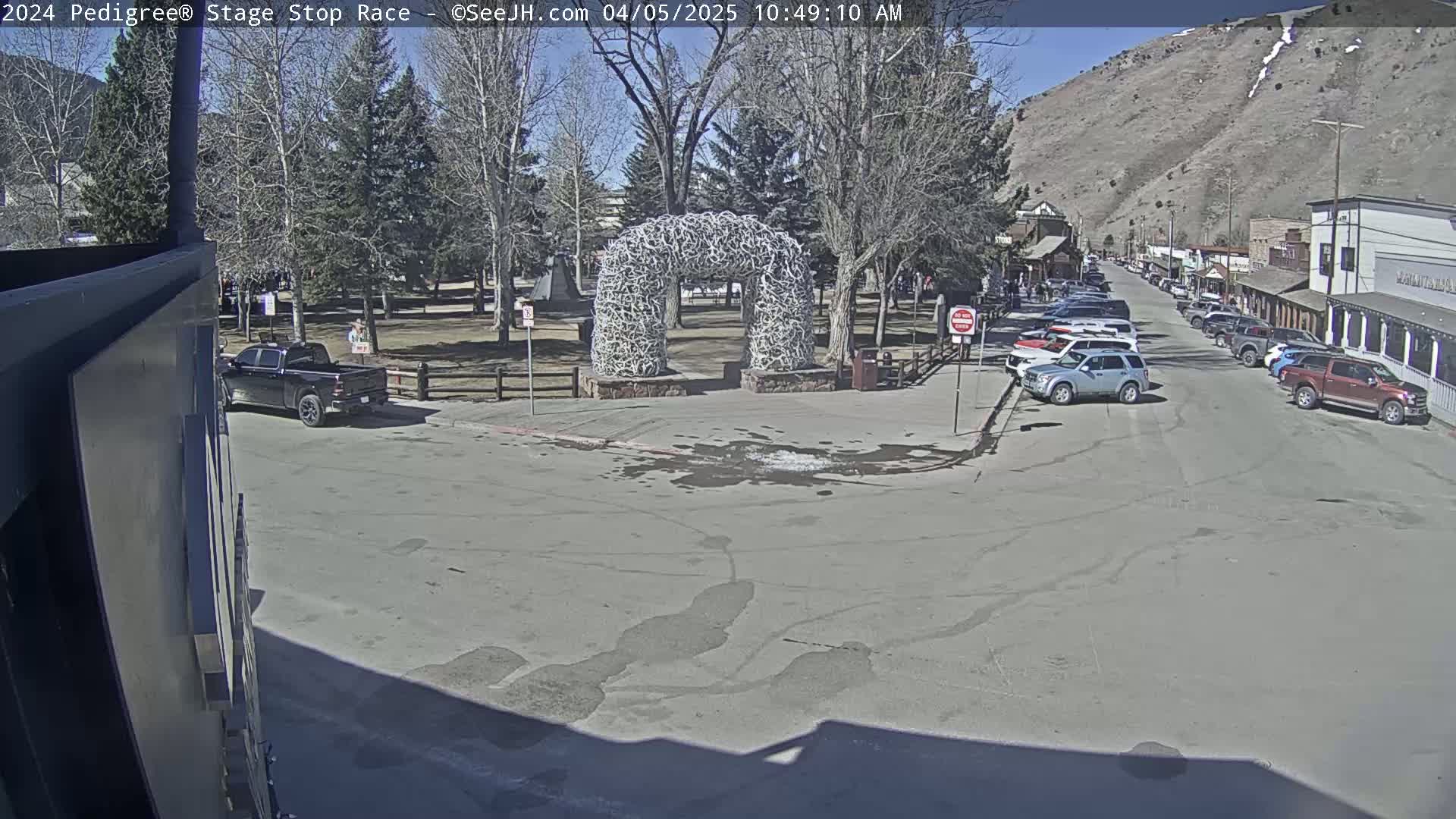 A sunny day in a town square features a large antler archway, several parked cars, and a mountain in the background.