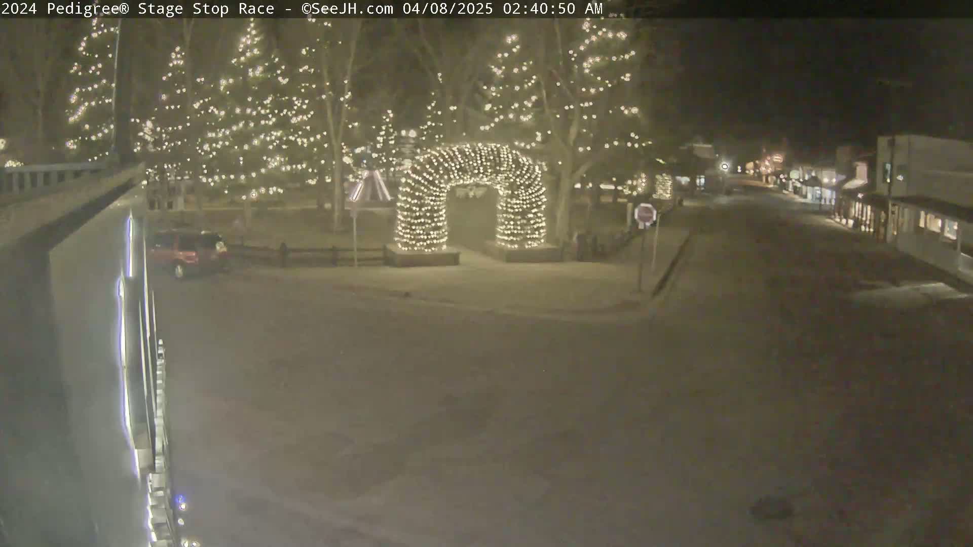 A snow-covered town square at night, illuminated by Christmas lights on trees and an archway, with a street visible in the background.