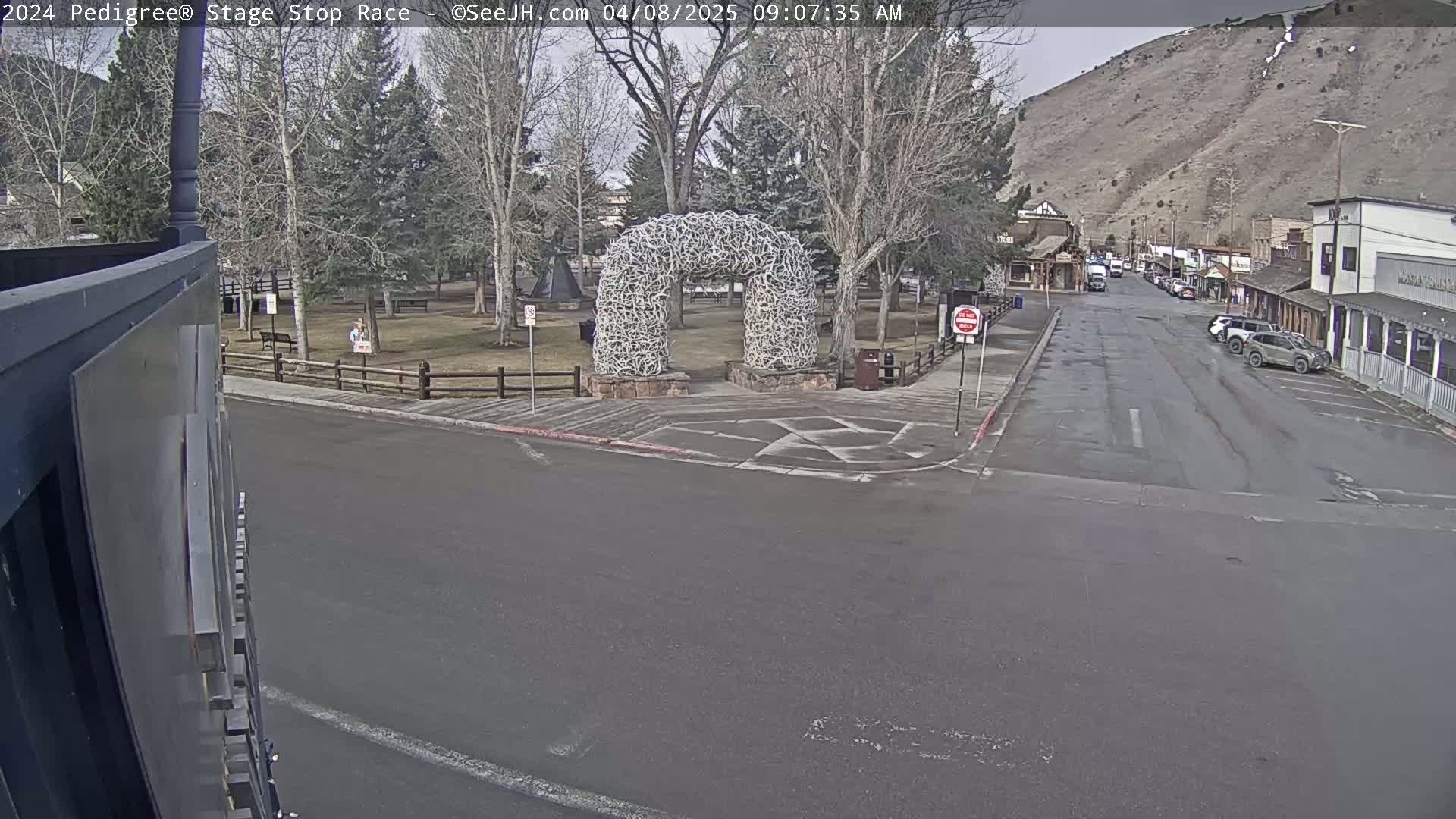 A town square with a large arch made of antlers is seen on an overcast day, with parked cars lining a street leading toward a mountain in the background.