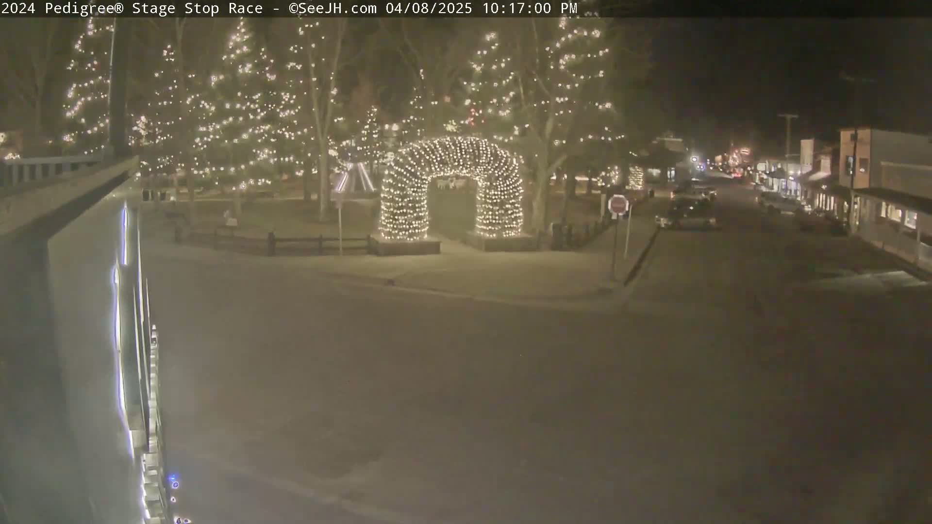 A nighttime scene shows a snow-covered town square with a lighted archway, Christmas trees decorated with lights, and cars parked along a street.