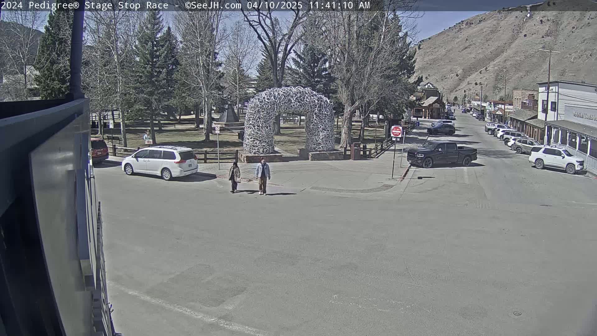 A sunny day shows a town square with a large antler arch, several parked cars, and two pedestrians walking.