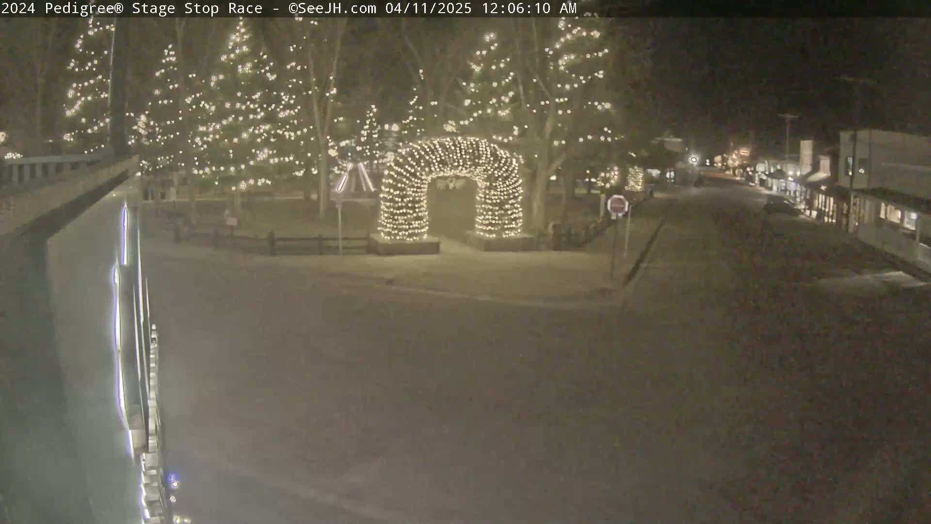 A snow-covered town square at night, illuminated by string lights on trees and an archway, with a street visible in the background.