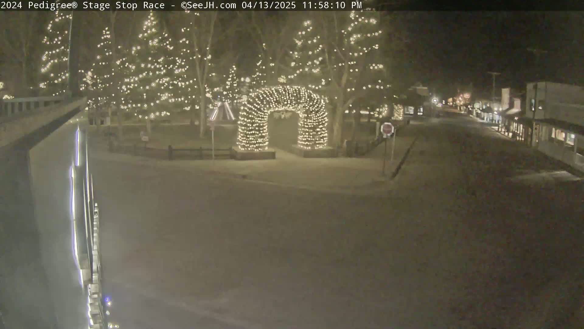 A snow-covered street at night, lined with buildings and trees decorated with Christmas lights, features a light-up archway in the center.