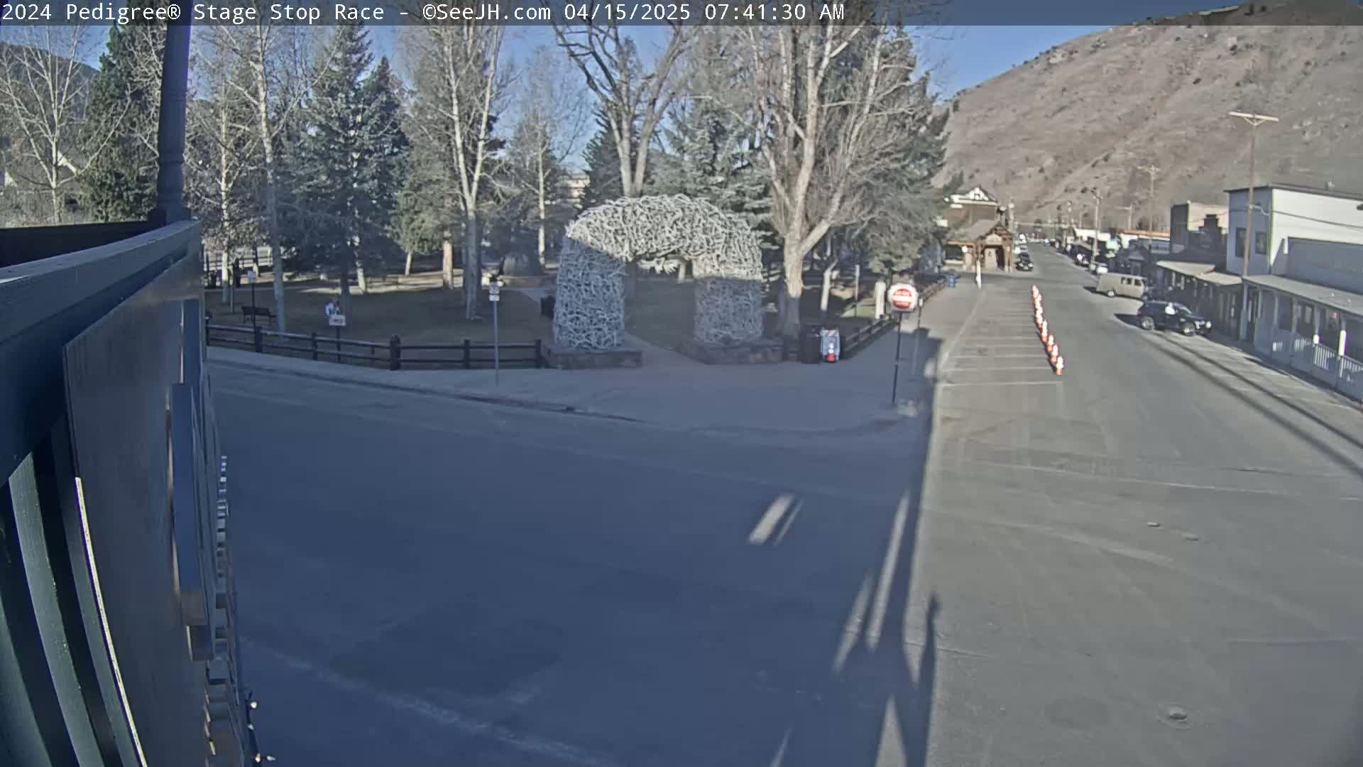 A sunny day shows a town street scene with a large antler archway in a park across the street from parked cars and buildings, and traffic cones lining the street.