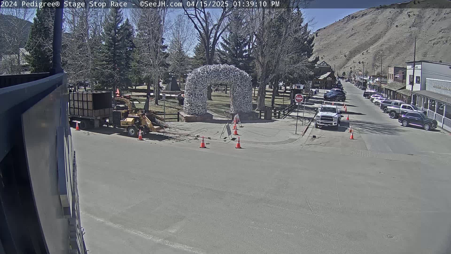 A sunny day shows a street scene with a large antler arch in the center, construction equipment, and parked cars lining a street leading toward buildings and a mountainside.