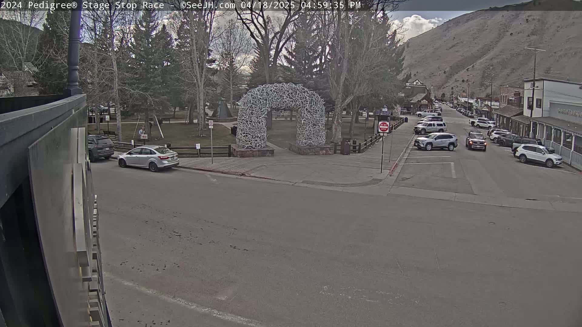 A mostly sunny day shows several cars parked along a street near a park with a large antler arch, with a mountain in the background.