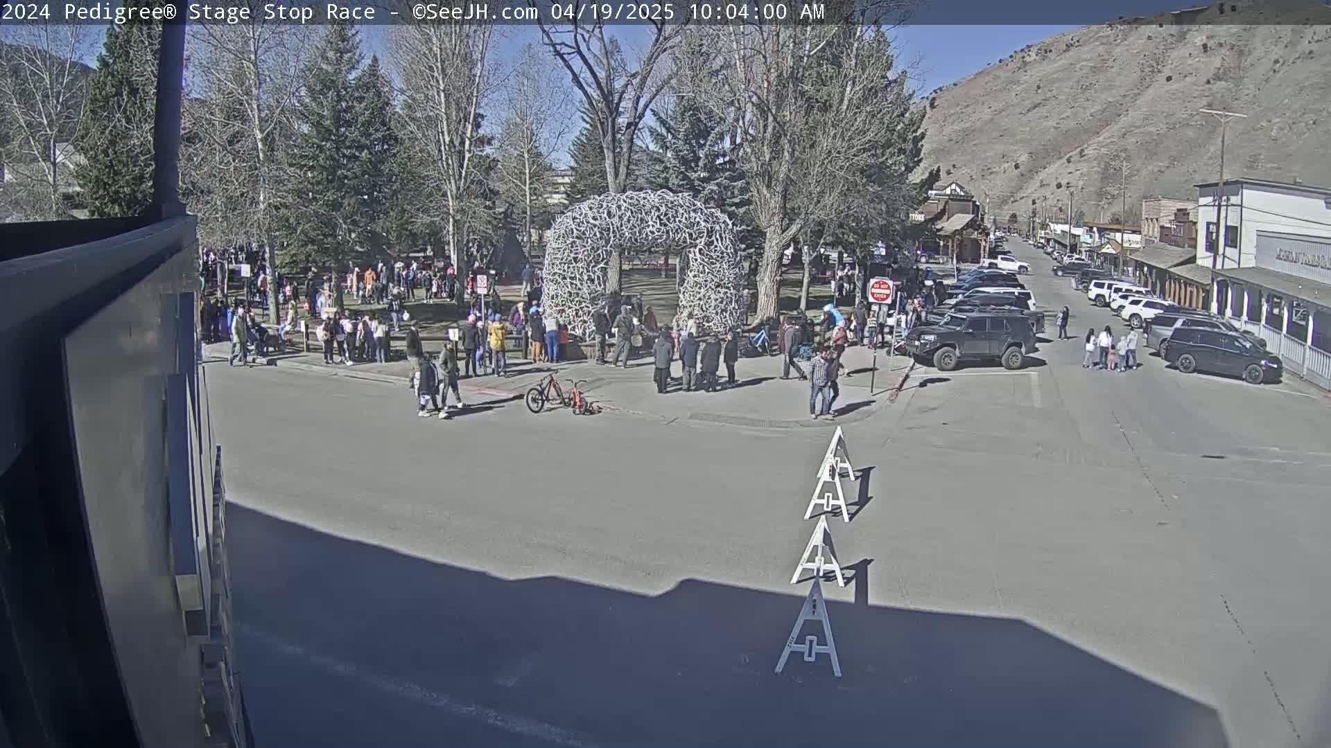 A sunny day in a town square shows a crowd of people gathered around a large antler arch, with parked cars lining the street and mountains in the background.