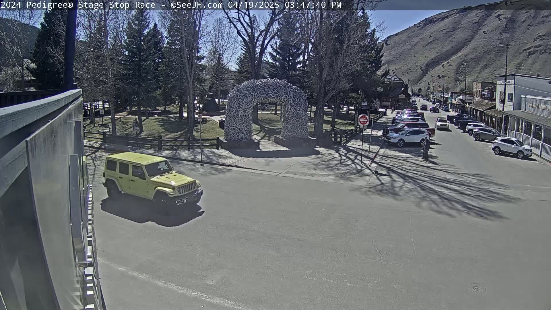 A yellow Jeep is driving through a town square on a sunny day, near a large arch made of antlers.