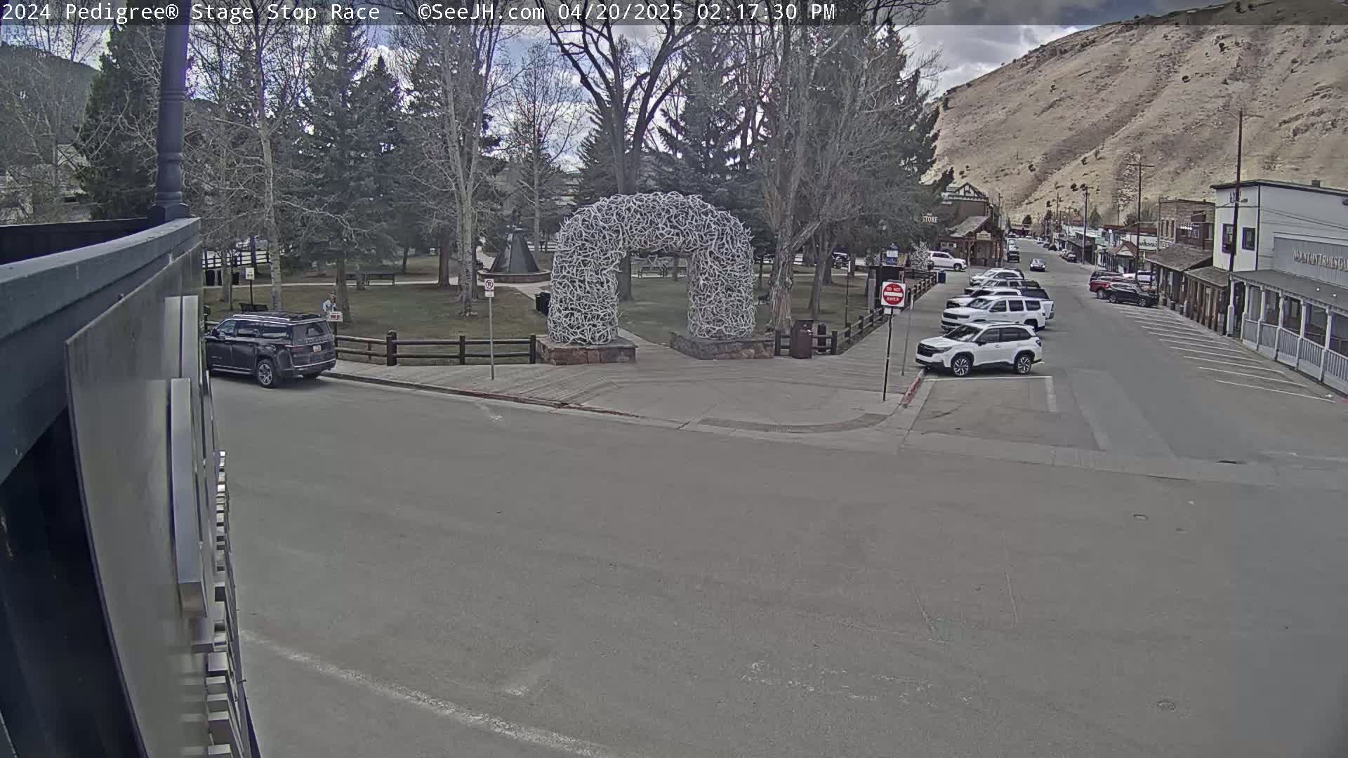 A partly cloudy day shows a street scene with parked cars, a large antler archway in a park-like setting, and a mountain in the background.