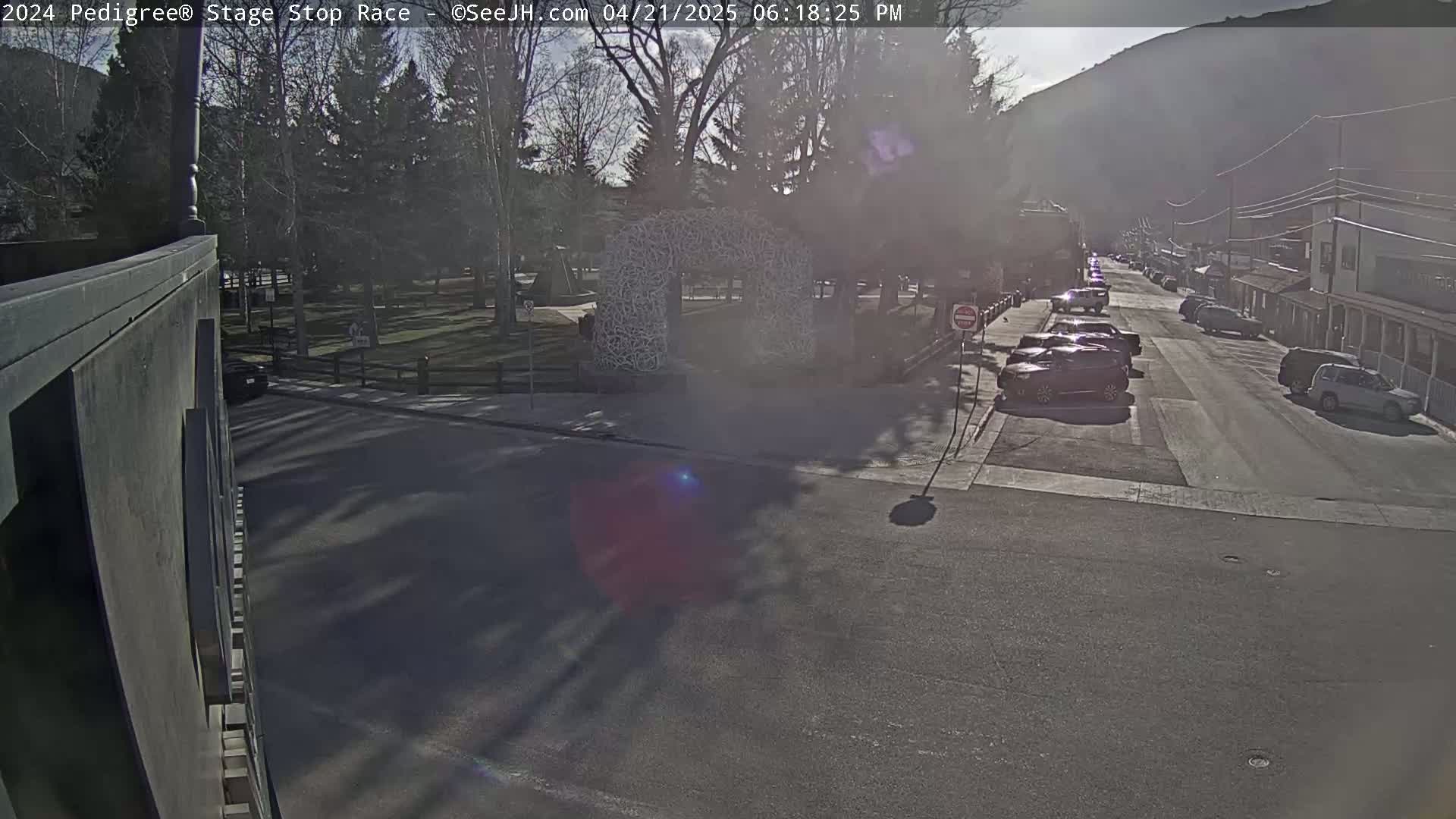 A sunny, hazy day reveals a street scene with parked cars lining a road that leads toward buildings nestled at the base of a mountain,  a park with a distinctive arch-like structure is visible in the foreground.