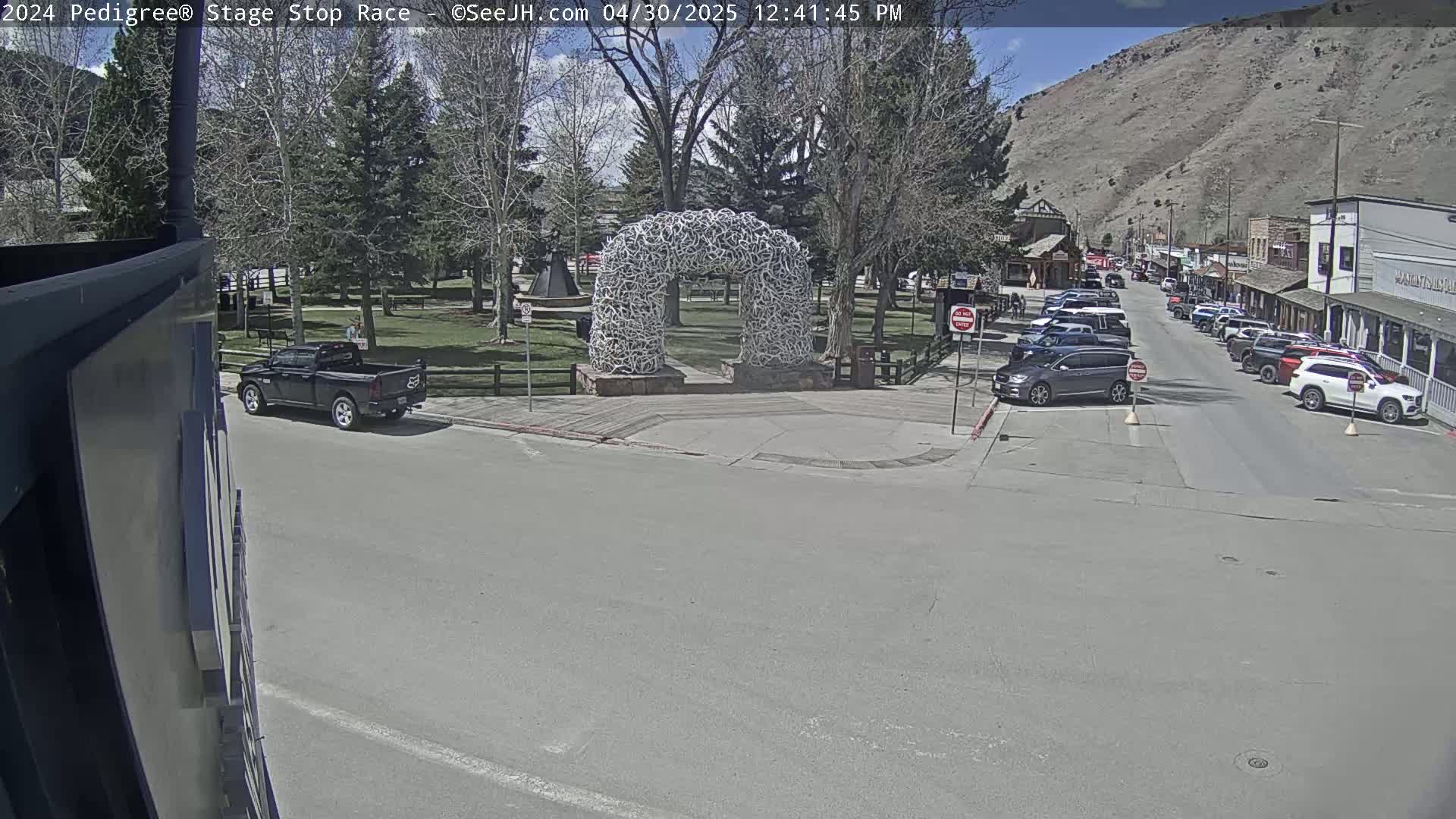 A sunny day reveals a town square with a large arch made of antlers, parked cars lining a street, and a pickup truck parked at the corner of a street.