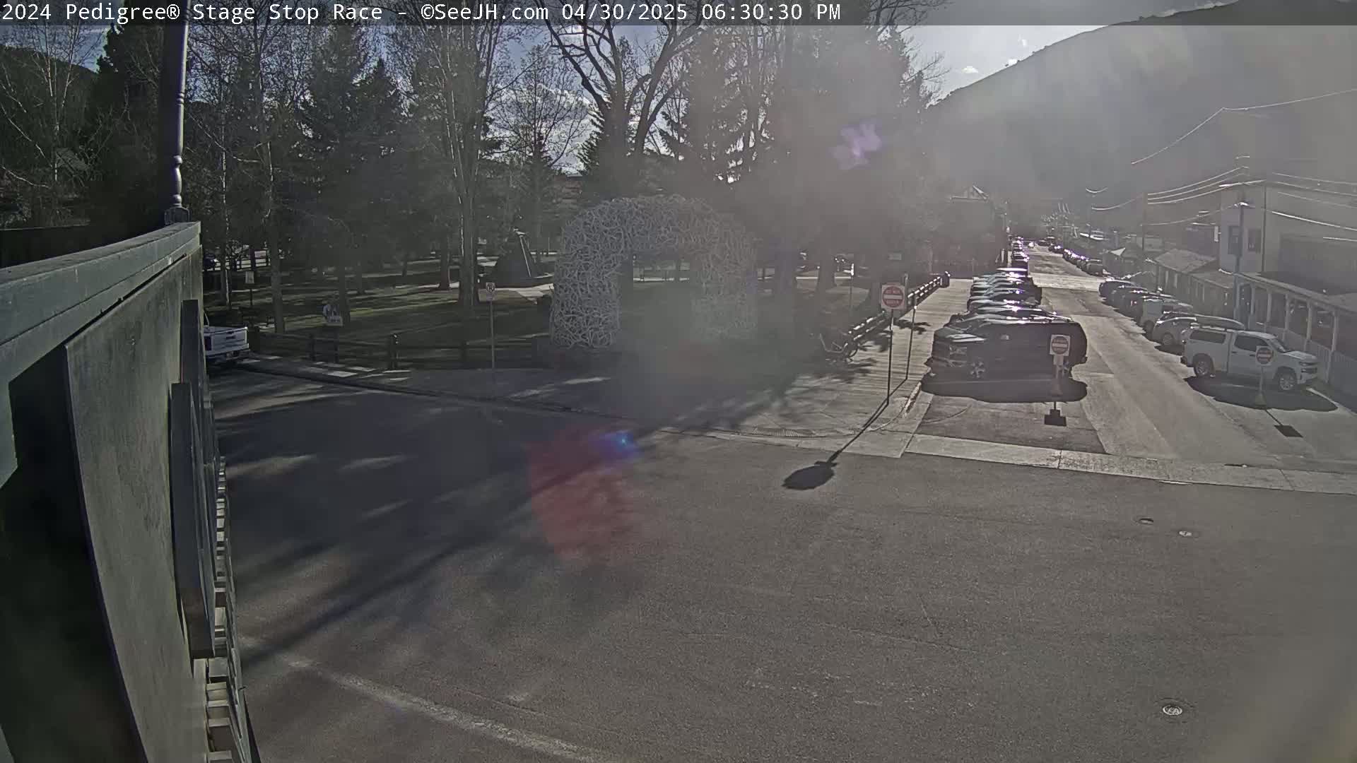 A sunny, hazy day reveals a street scene with parked cars lining the sides, a park with trees across the street, and a mountain in the background.