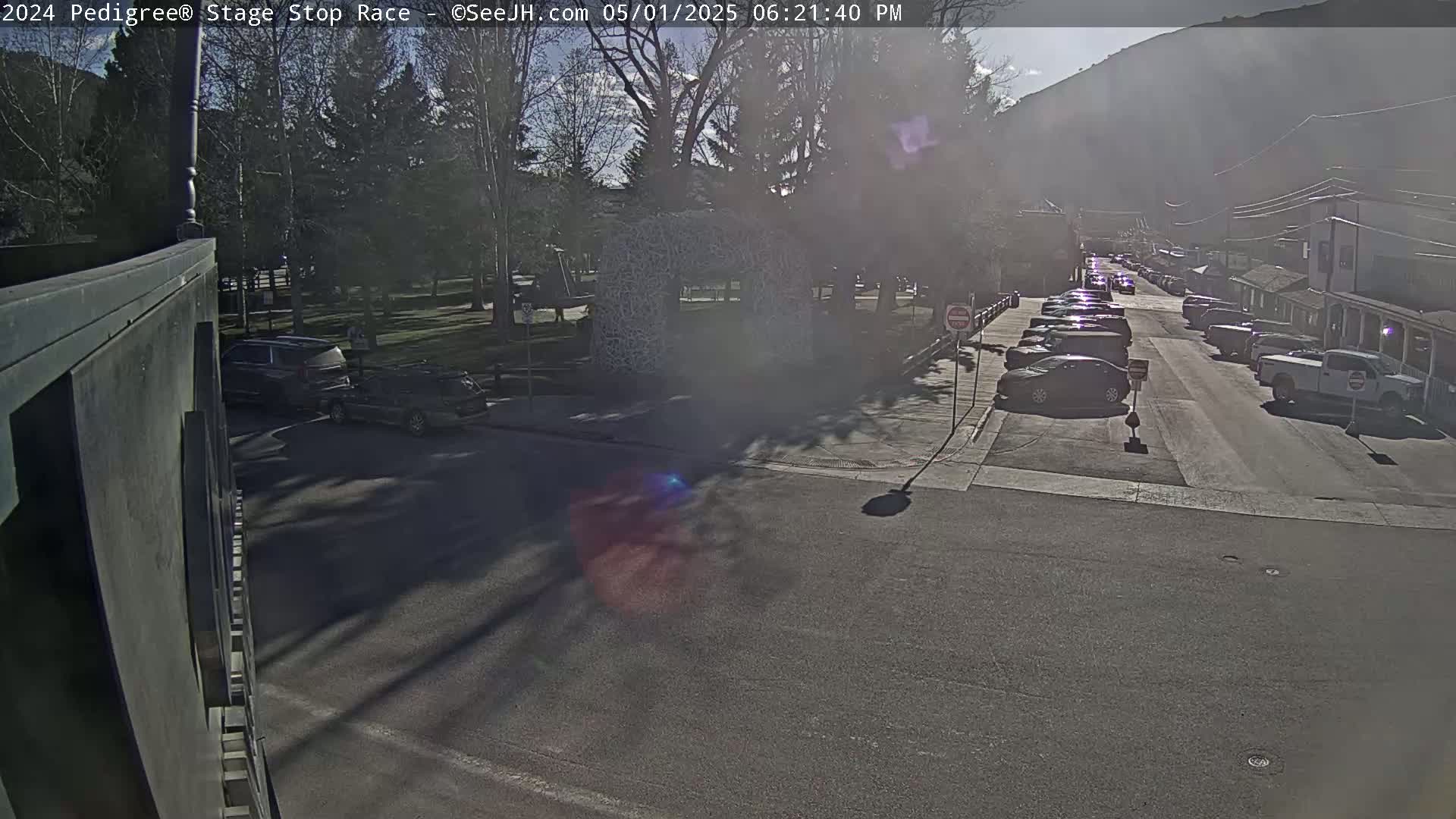 A sunny day reveals a street scene with parked cars lining both sides of a road, leading towards buildings nestled against a mountainside.
