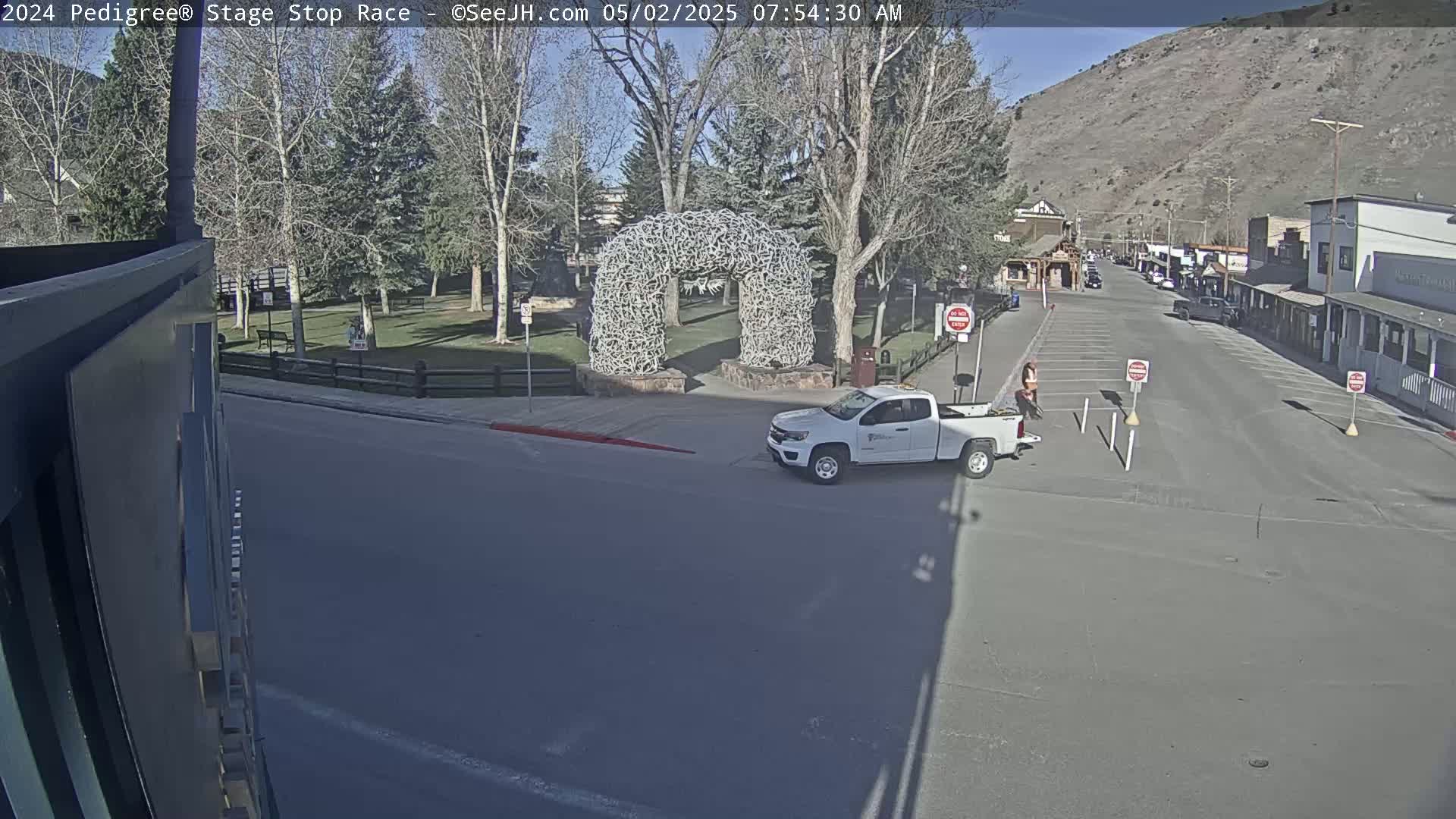 A sunny day shows a white pickup truck parked near a large arch made of antlers in a town with a mountain in the background.