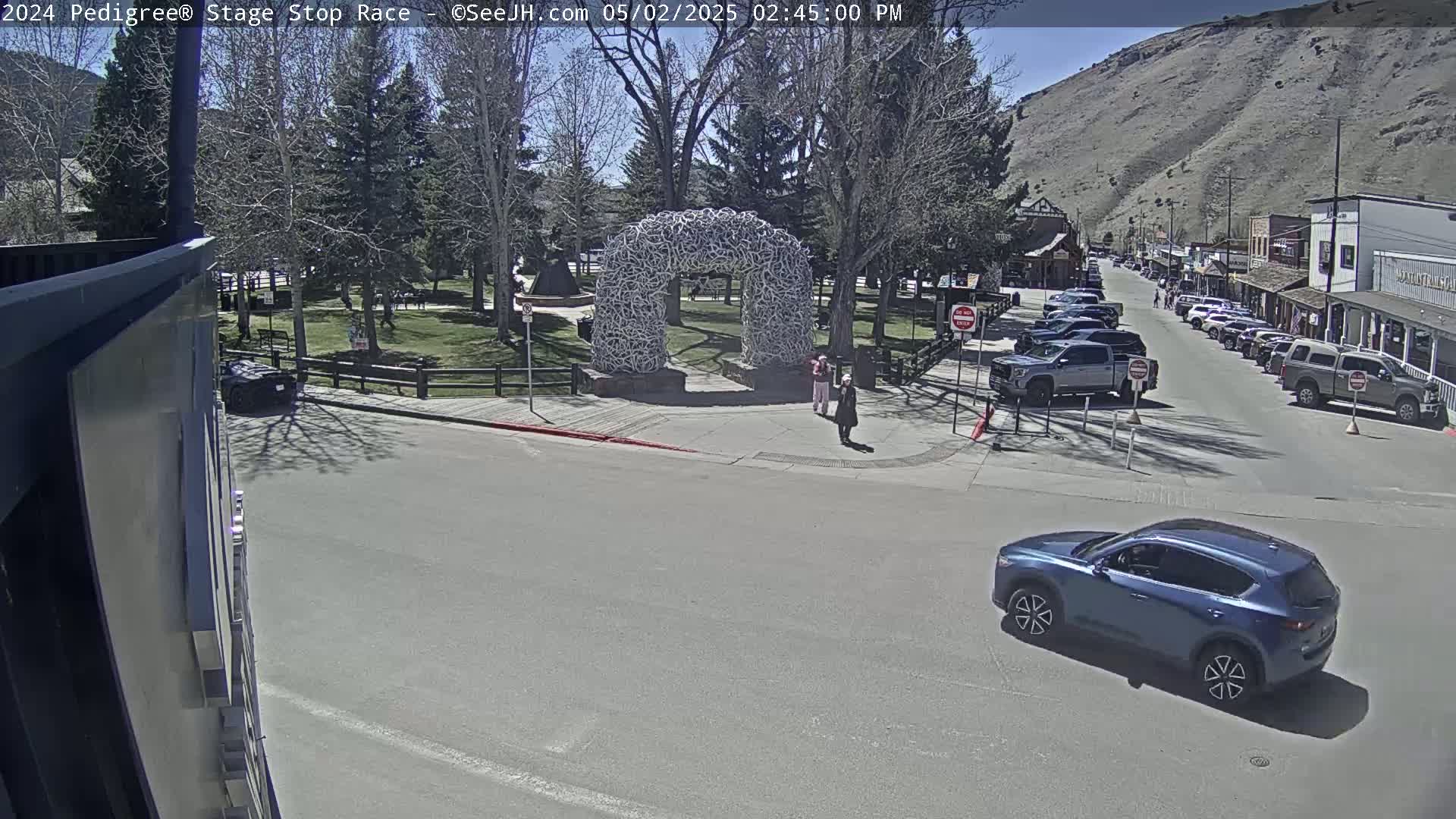 A blue SUV drives past a town square on a sunny day, near an arch made of antlers.