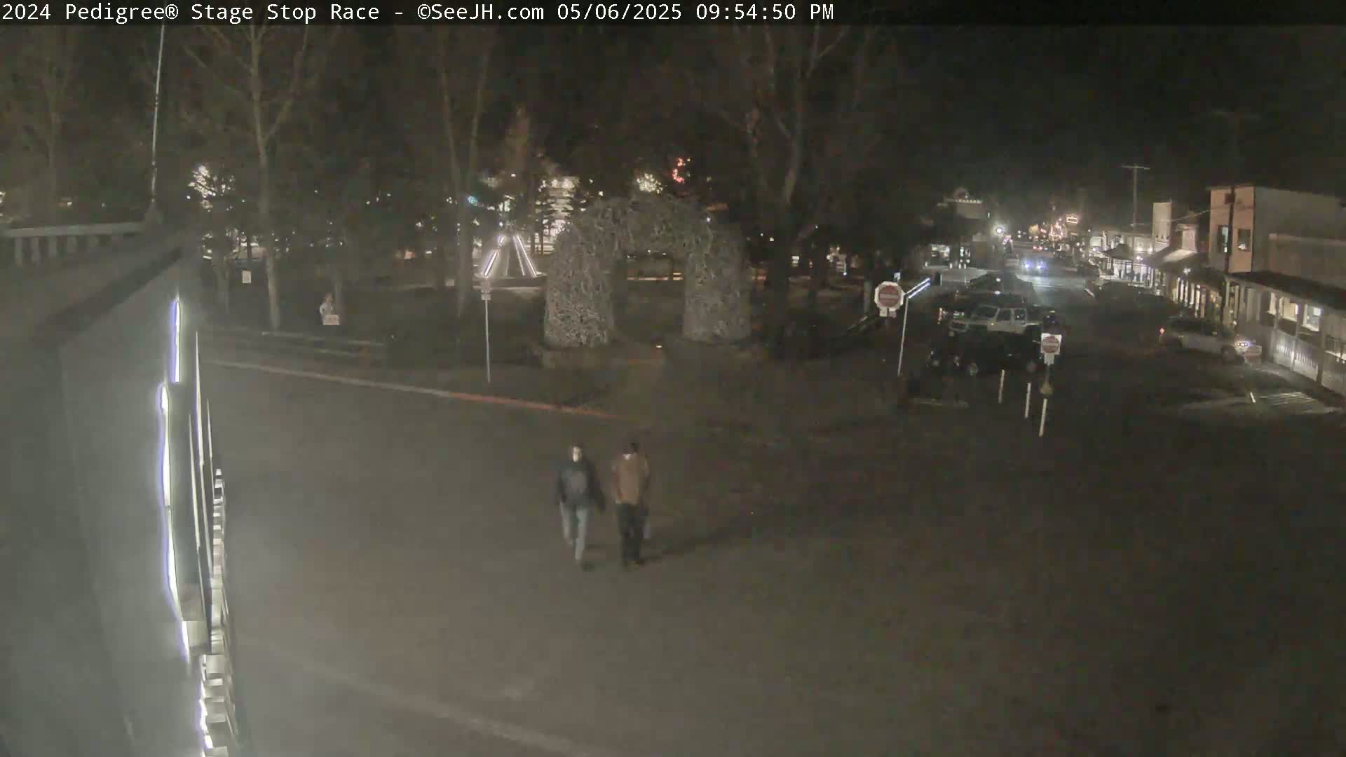 Two people walk across a dark street at night near a town square with decorative structures, under mostly clear skies.