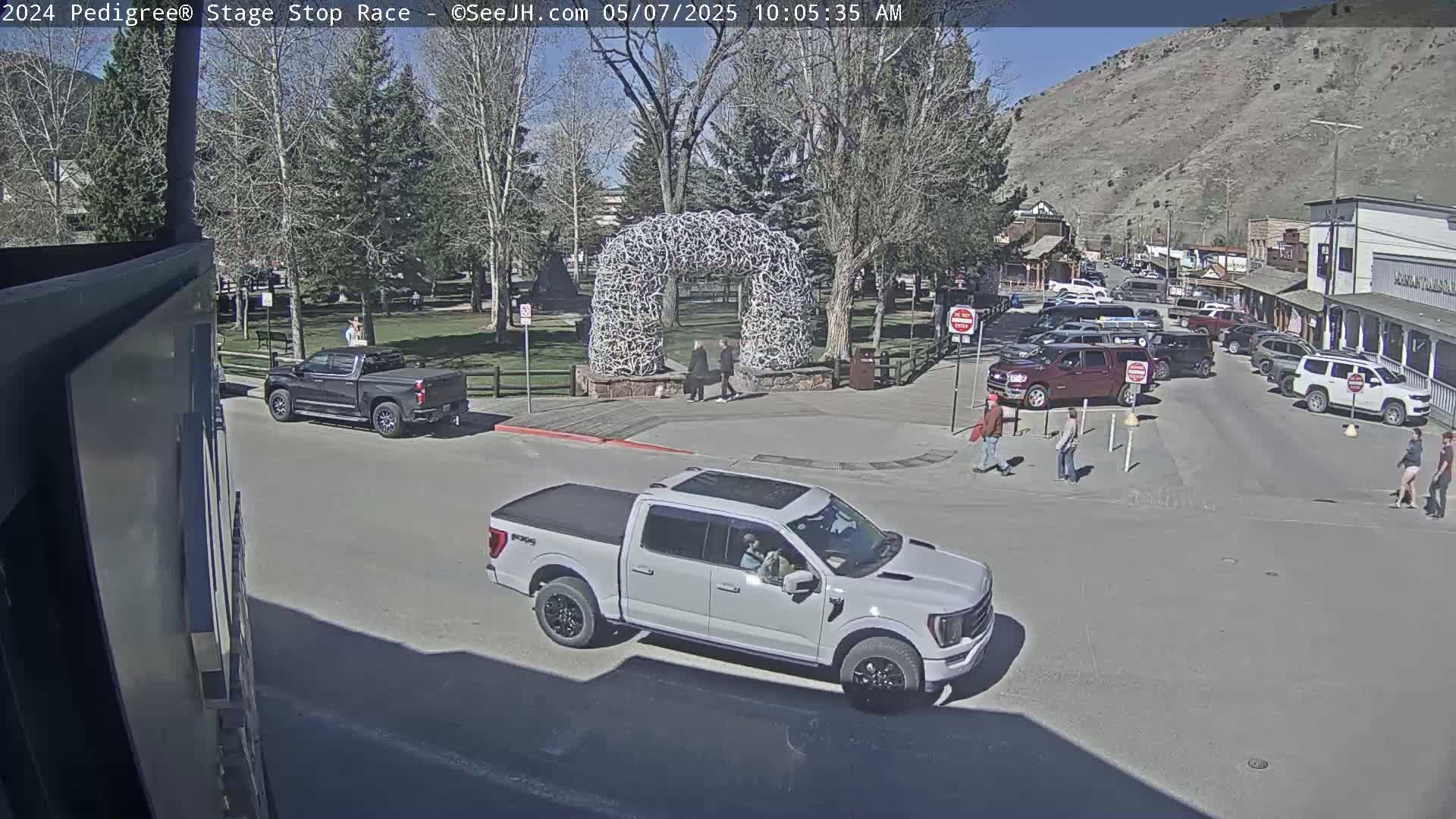 A sunny day shows a town square with a large antler arch, several vehicles, and pedestrians walking on a street.