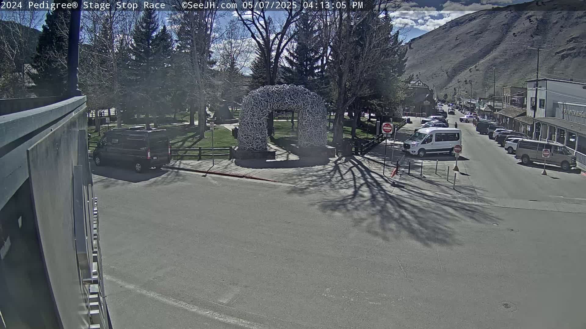 A sunny day shows a town square with a large arch made of antlers, several parked vehicles, and a street with cars driving on it, all situated against a mountainous backdrop.