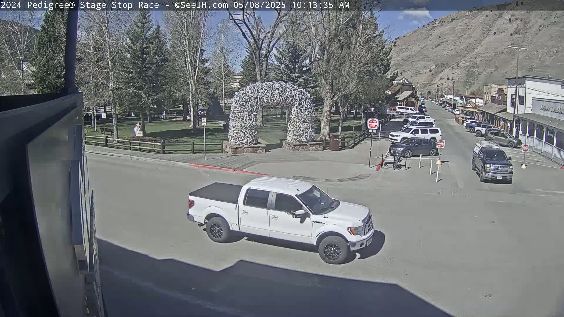 A white pickup truck is parked near a street corner, next to a park with a large arch made of antlers under a sunny sky.