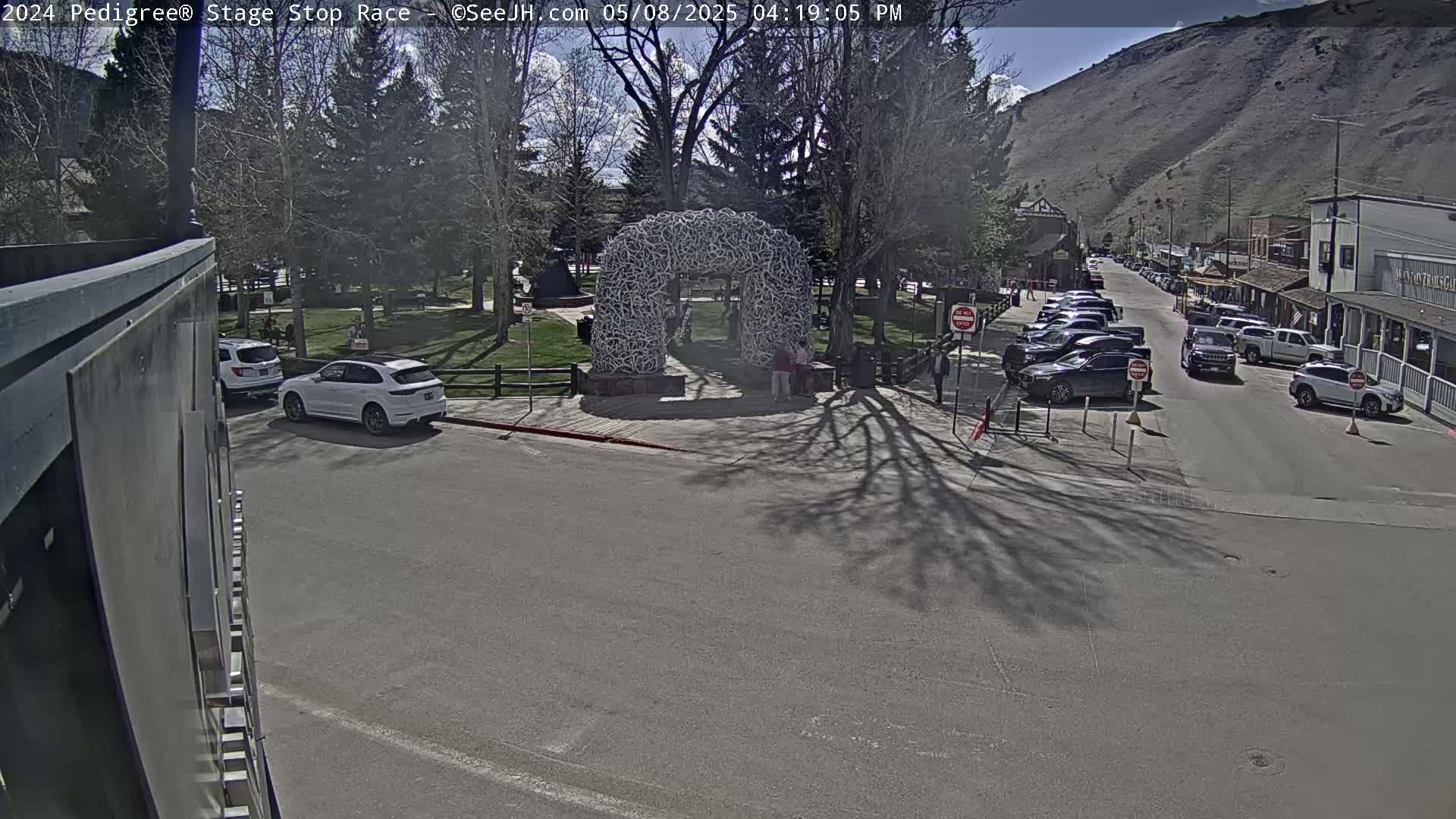 A sunny day shows a town street with parked cars, a park with a large antler arch, and a mountain in the background.