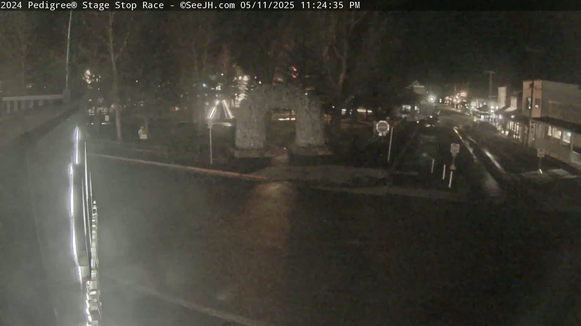 A nighttime, wet street scene shows a stone archway in a park-like setting, with buildings and vehicles visible in the distance.