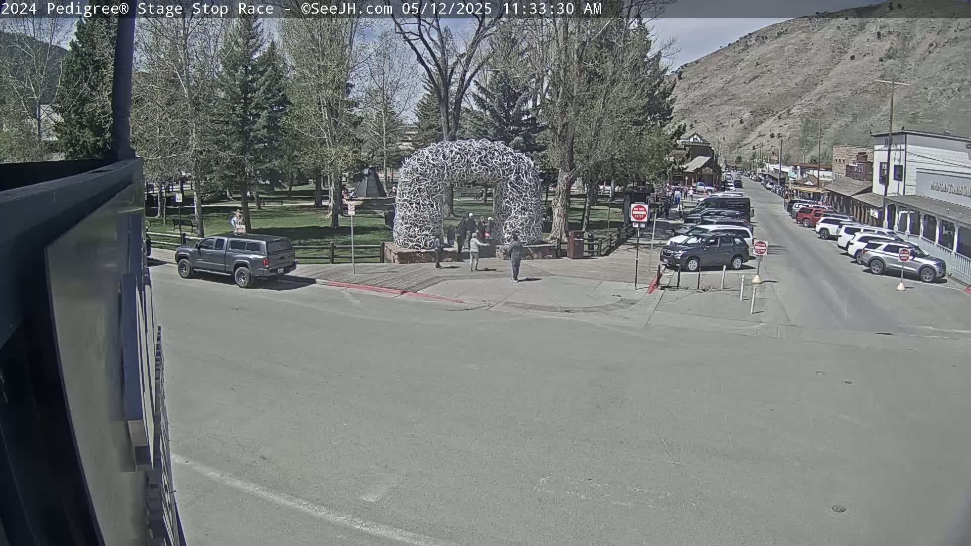A sunny day reveals a town square with a large antler archway, several people standing near it, cars parked along a street lined with buildings, and a truck driving away from the arch.