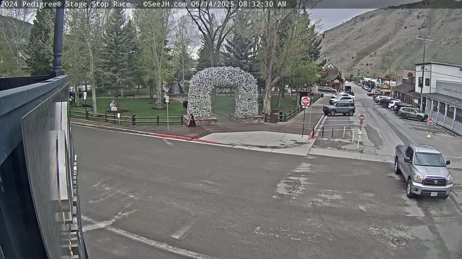 A street scene shows a silver pickup truck turning a corner near a park with a large arch-shaped sculpture made of antler-like materials, under an overcast sky.