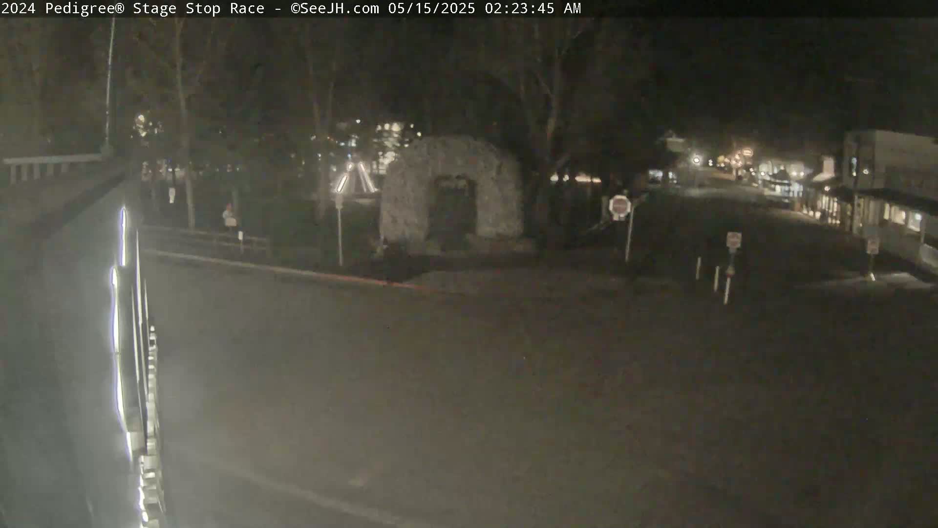 A nighttime, outdoor view of a mostly empty street with a stone archway structure in the center, surrounded by trees and buildings.