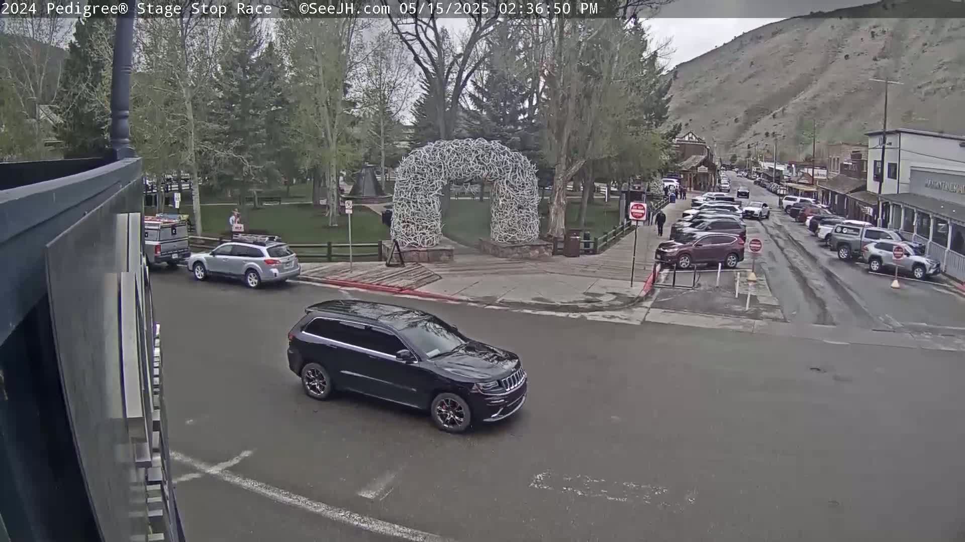 A dark-colored SUV drives past an antler arch in a town square under an overcast sky, with several parked cars lining a street of buildings on a hillside.