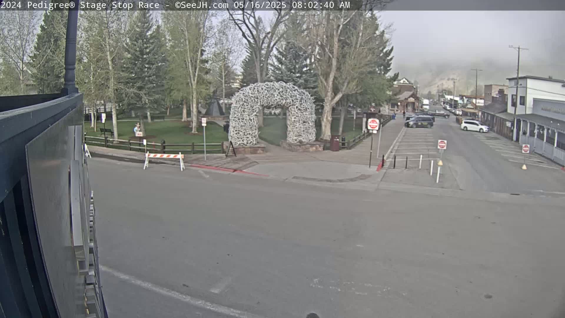 A foggy morning scene shows a town street with parked cars, a large antler archway in a park across the street, and a few people in the distance.