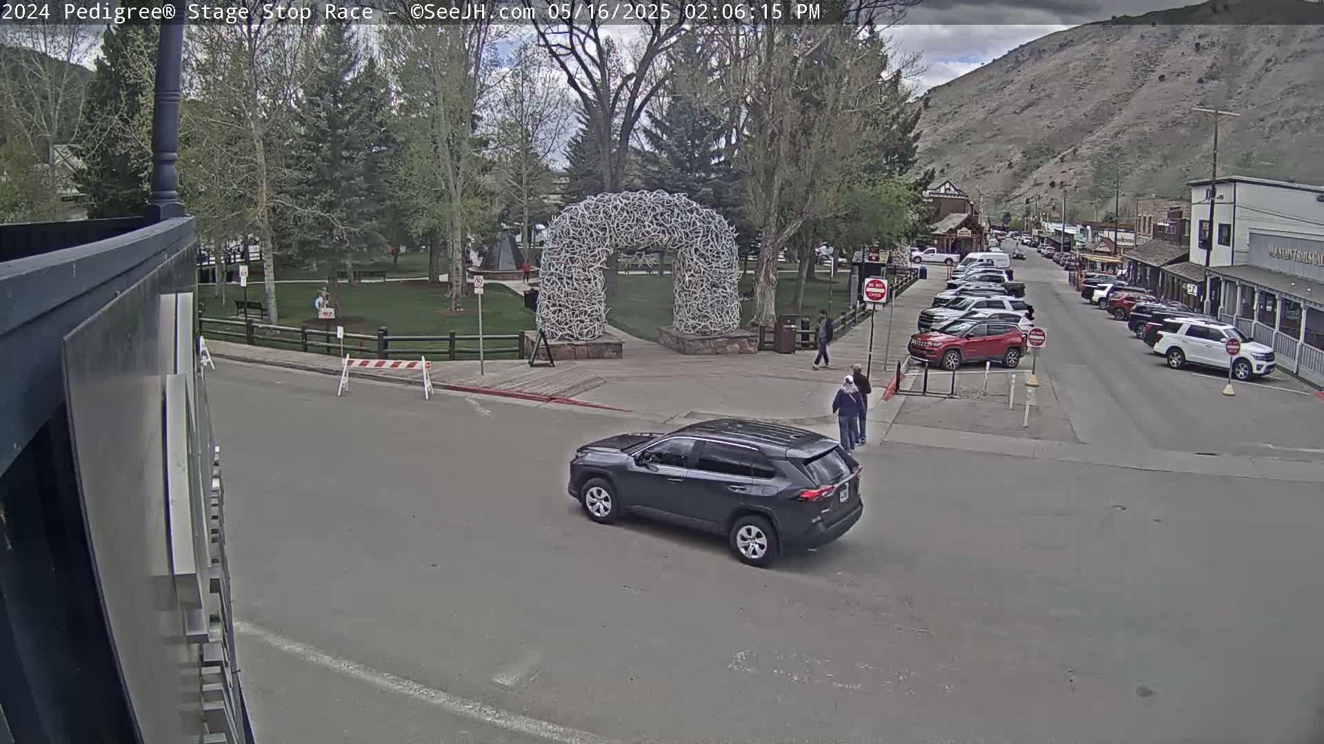 A dark-colored SUV is driving past a large antler archway in a town square on a partly cloudy day, with parked cars lining a street in the background and mountains visible in the distance.