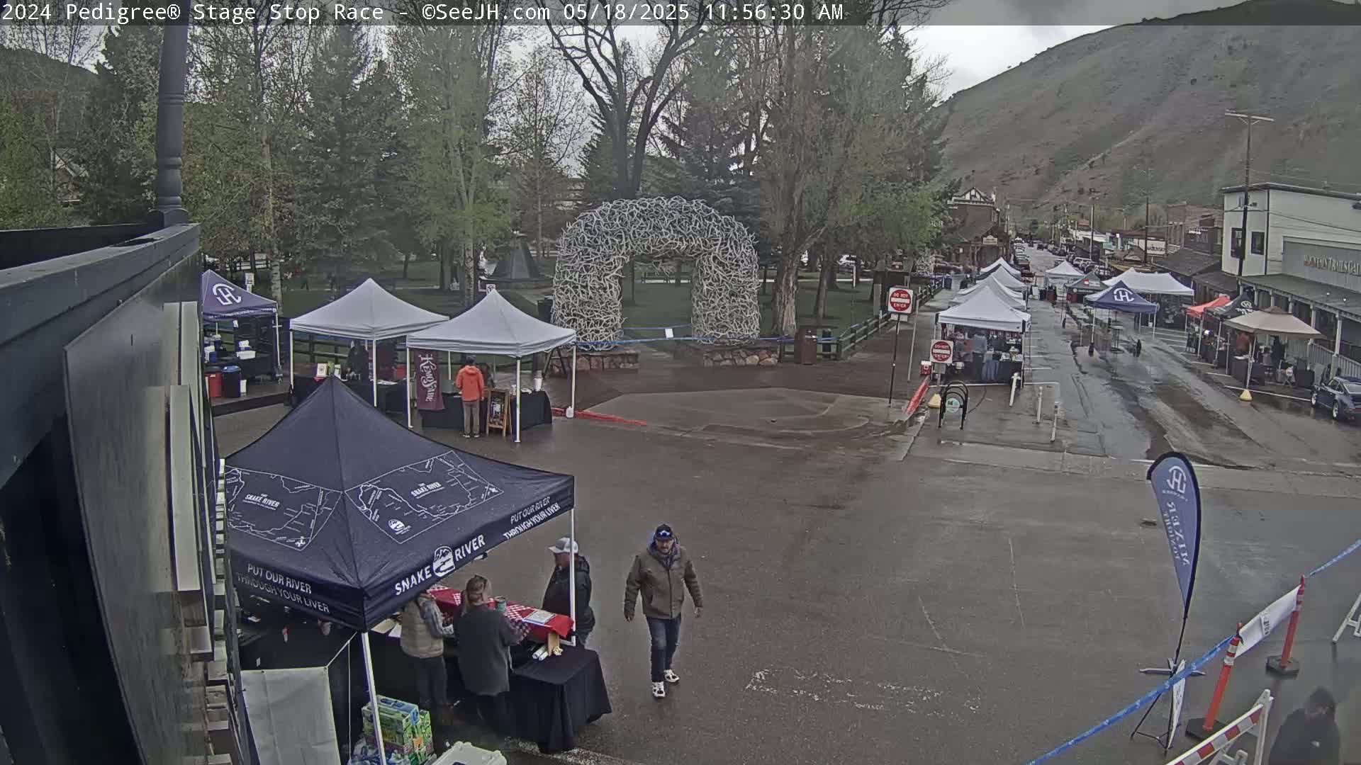 A wet town square with numerous vendor tents and a large antler archway under an overcast sky.