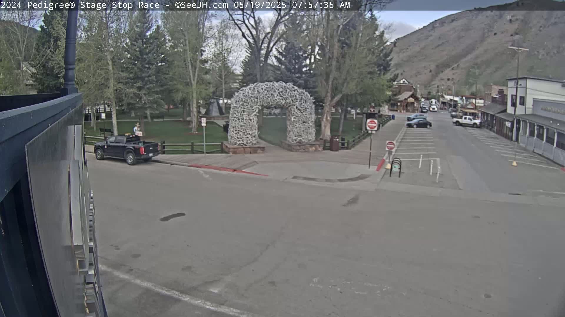 A mostly sunny day reveals a town square with a large arch made of antlers, a pickup truck parked nearby, and a few cars driving on a street that leads to buildings nestled against a mountainside.