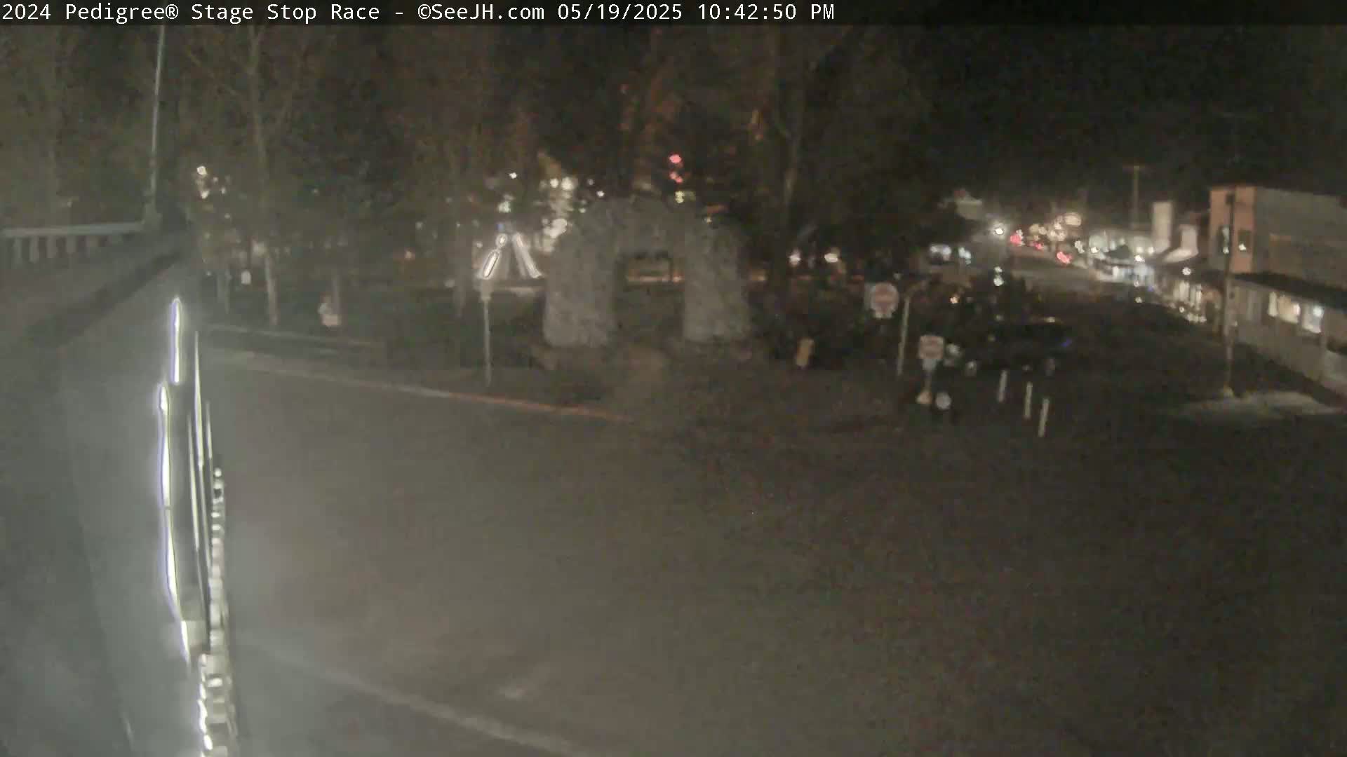 A nighttime, outdoor view shows a mostly empty street with a few cars and pedestrians near a stone archway under trees.