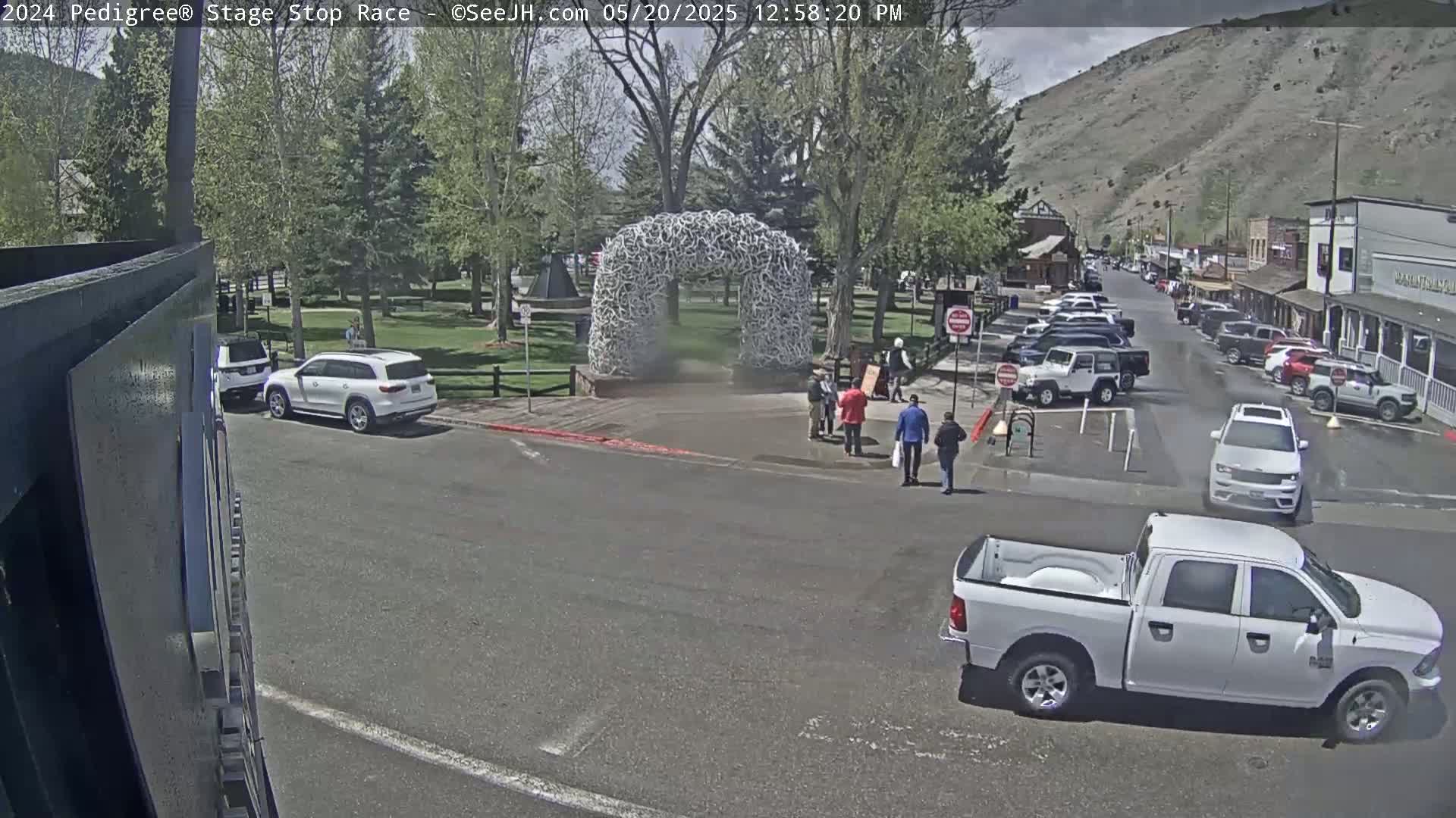 A sunny day shows a town square with a large antler arch, several parked cars, a few people walking, and a white pickup truck in the foreground.